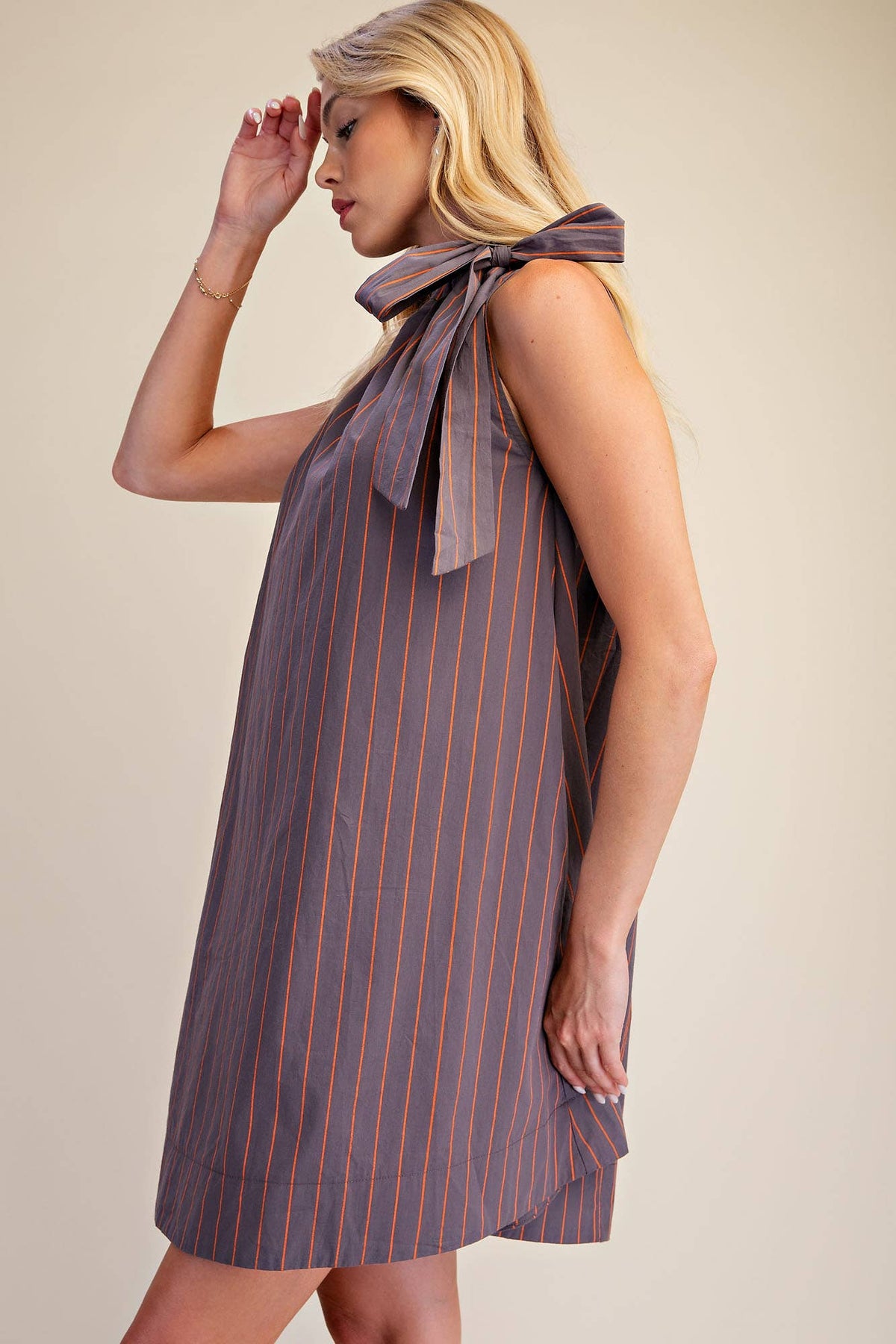 A woman wearing a sleeveless, high-neck shift dress with vertical orange pinstripes on a muted gray-purple base fabric, posed against a solid beige background.