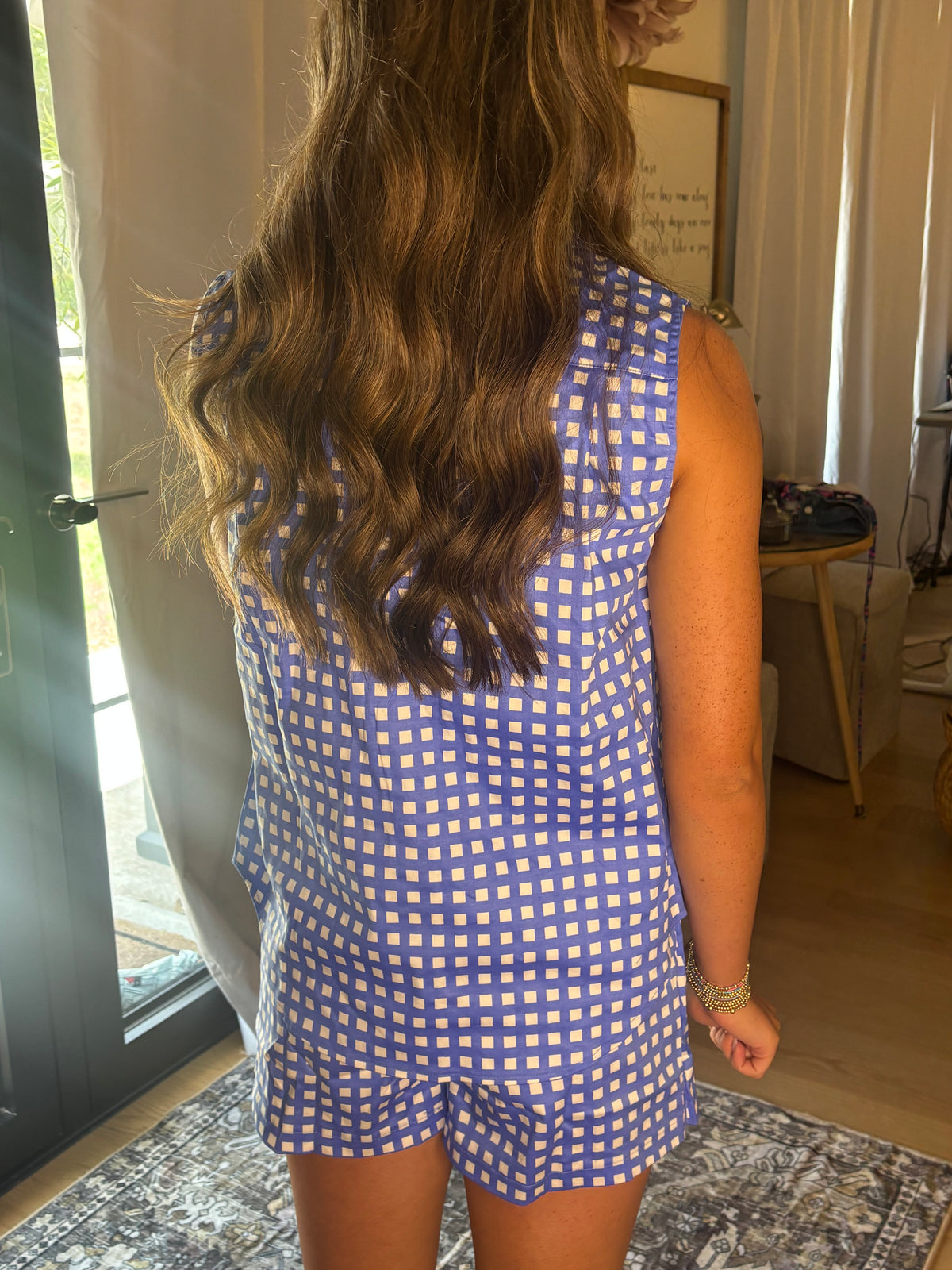 A woman viewed from behind wearing a sleeveless blue and white gingham top in a home setting with curtains and a wooden table in the background.