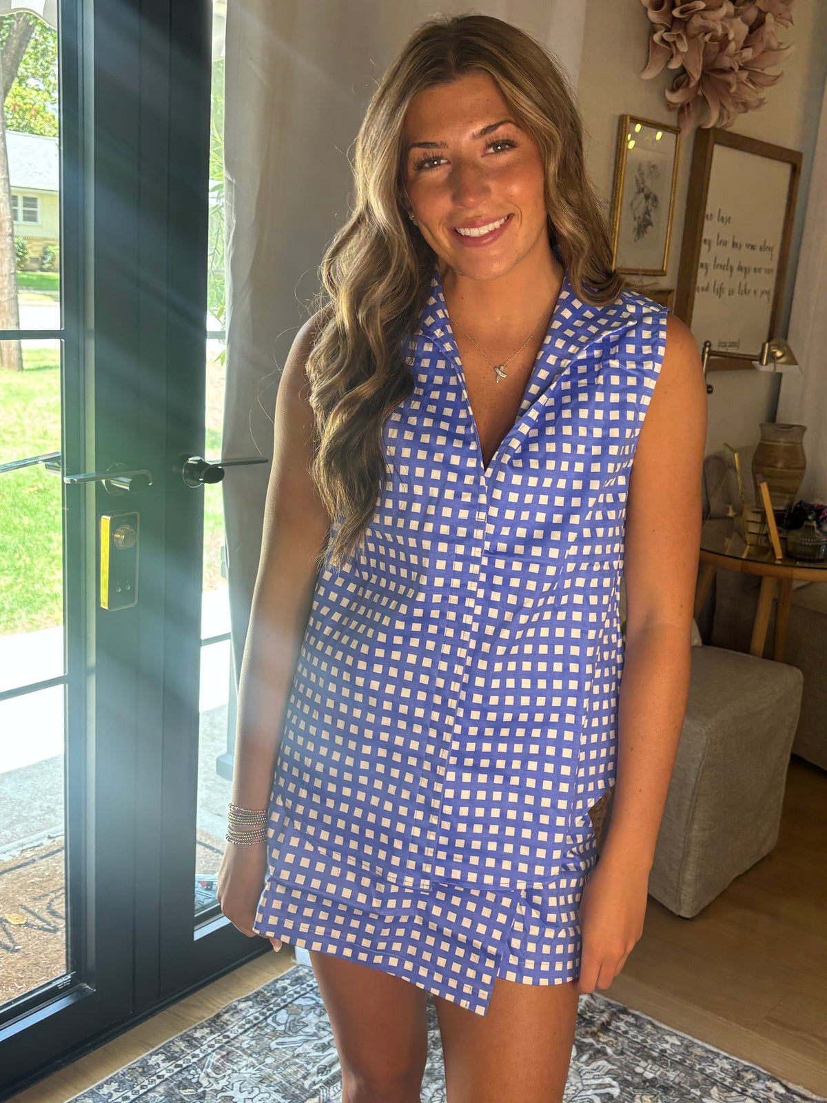 A woman wearing a sleeveless blue and white gingham top + skort, standing in a home interior near a glass door.