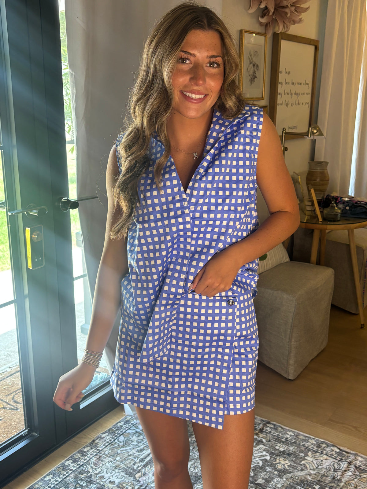 A woman wearing a blue and white gingham sleeveless collared top & skort, standing in a brightly lit home interior with a glass door, rug, and side table visible.