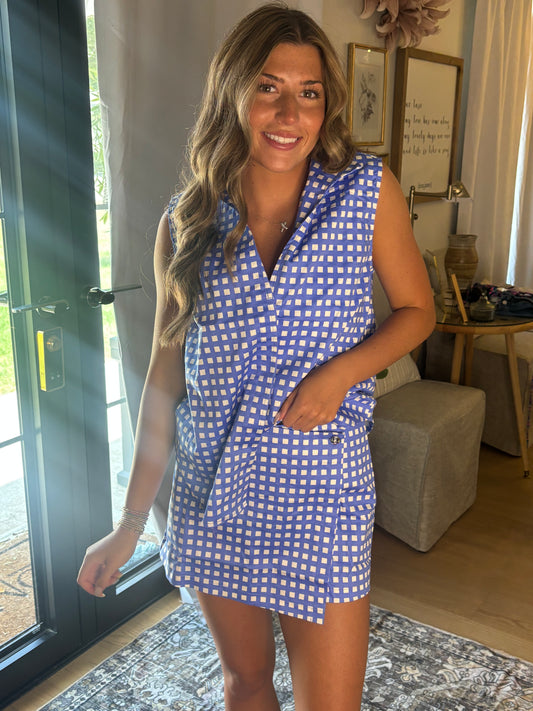 A woman wearing a blue and white gingham sleeveless collared top & skort, standing in a brightly lit home interior with a glass door, rug, and side table visible.