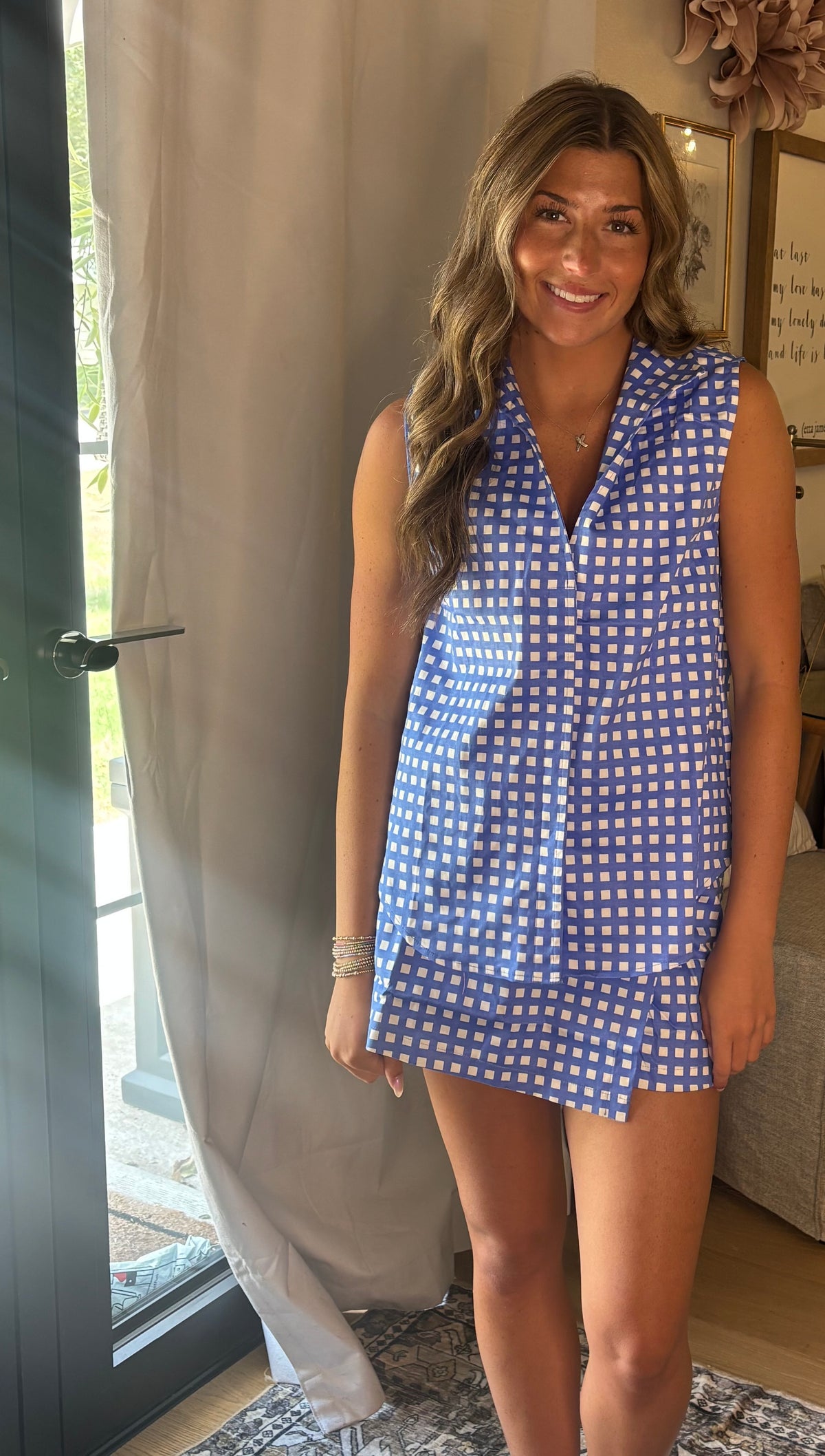 A woman wearing a sleeveless blue and white gingham top & skort standing in a home setting next to a glass door and curtain.