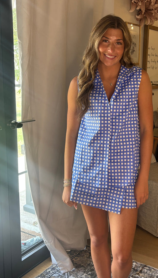A woman wearing a sleeveless blue and white gingham top & skort standing in a home setting next to a glass door and curtain.