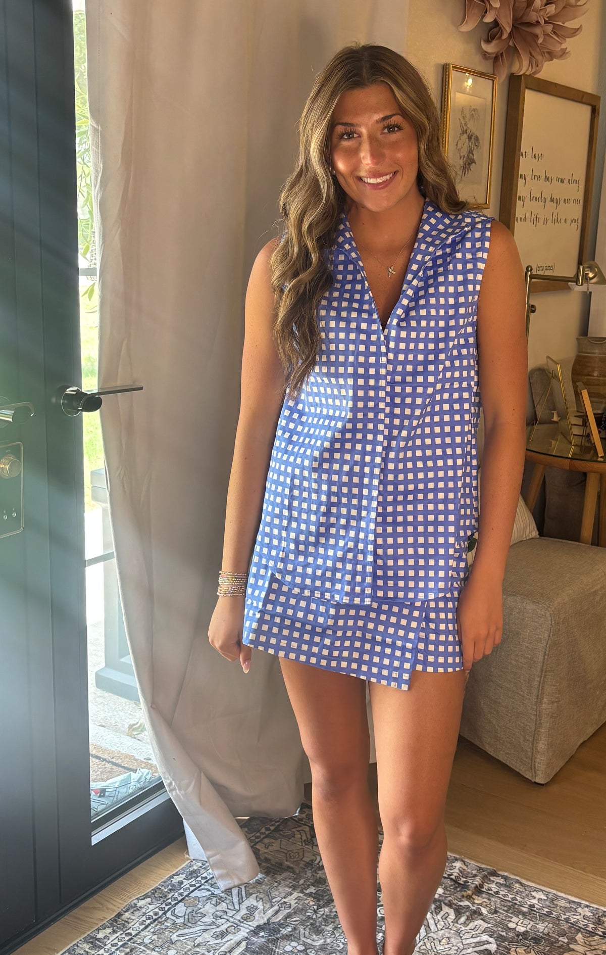 A woman modeling a sleeveless blue and white gingham top and skort in a home setting with curtains, a door, and a rug visible.