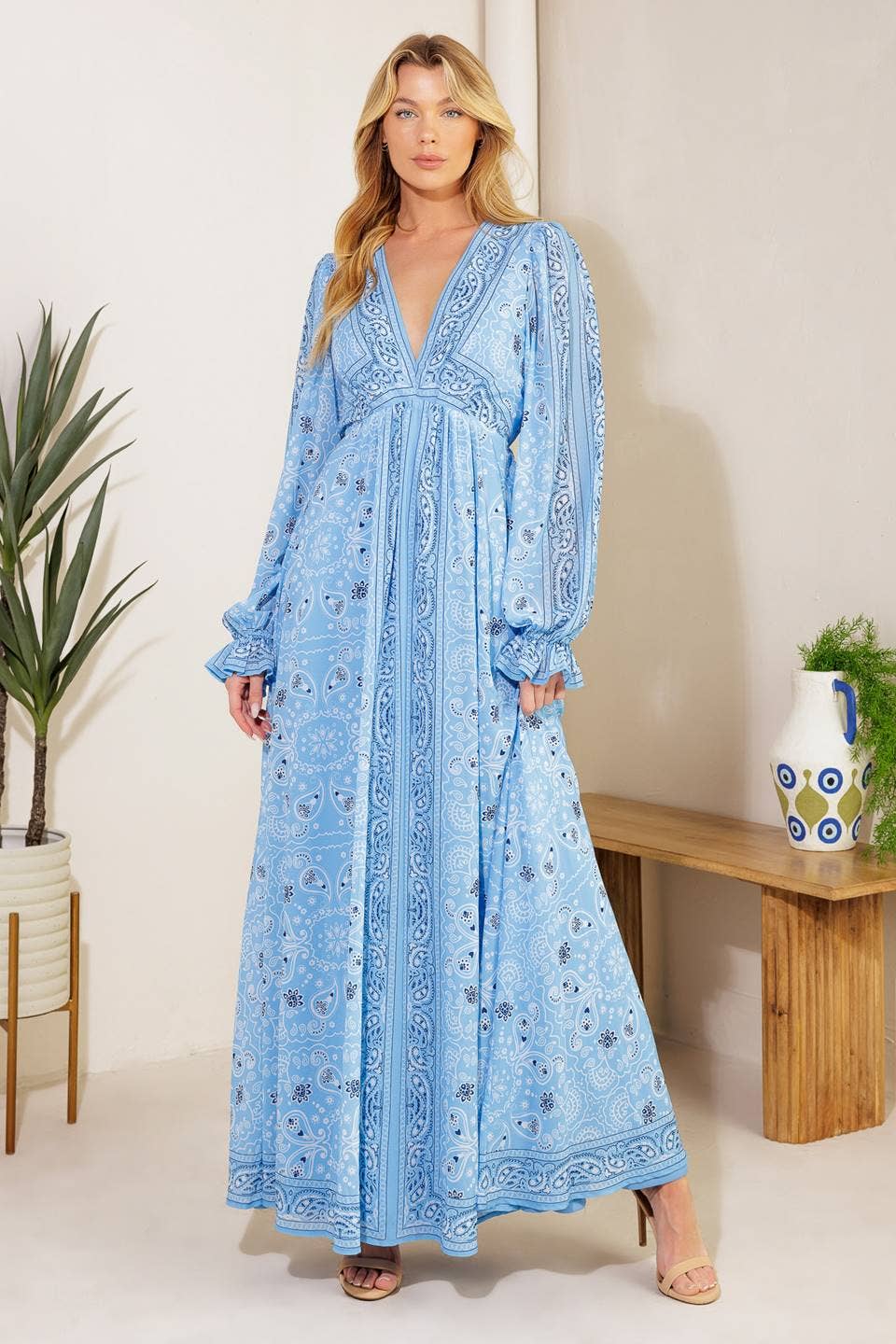 Woman wearing a blue patterned dress in a room with plants and a wooden bench.