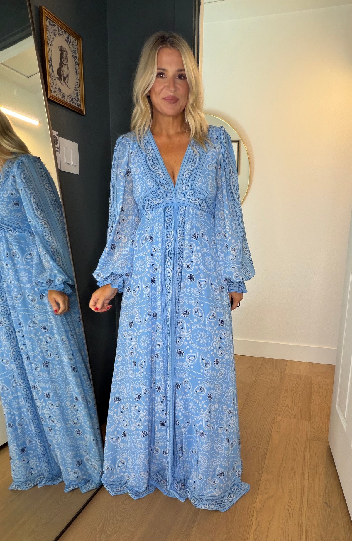 A woman wearing a light blue bohemian-style maxi dress with a floral print, standing in front of a mirror that reflects her side profile. The setting is a home interior with light wood floors, white walls, and a dark gray accent wall.