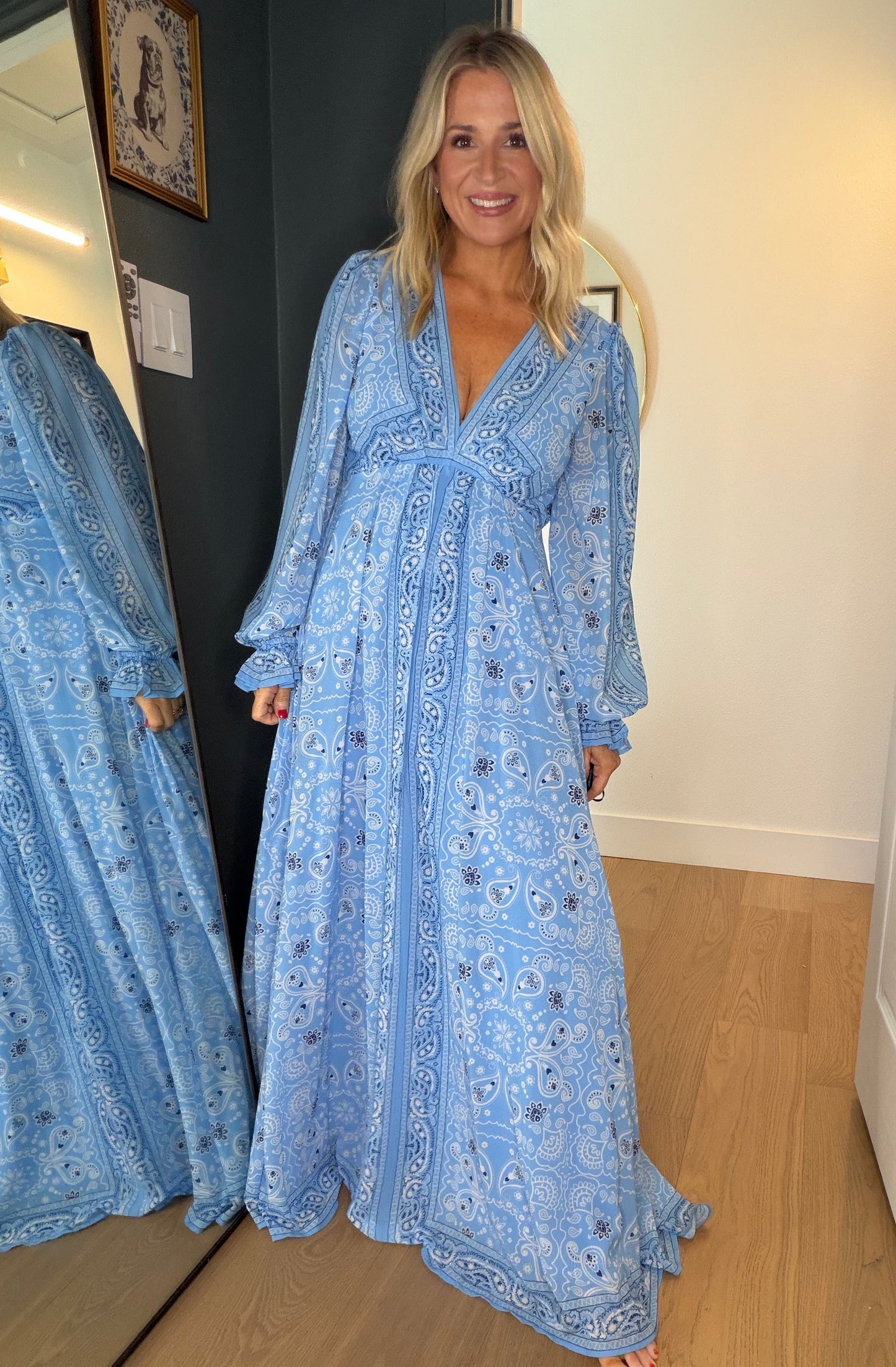 A woman wearing a light blue patterned maxi dress stands in a doorway, with her reflection visible in a mirror on the left. The dress has a deep V-neck, long sleeves, and a flowing silhouette.