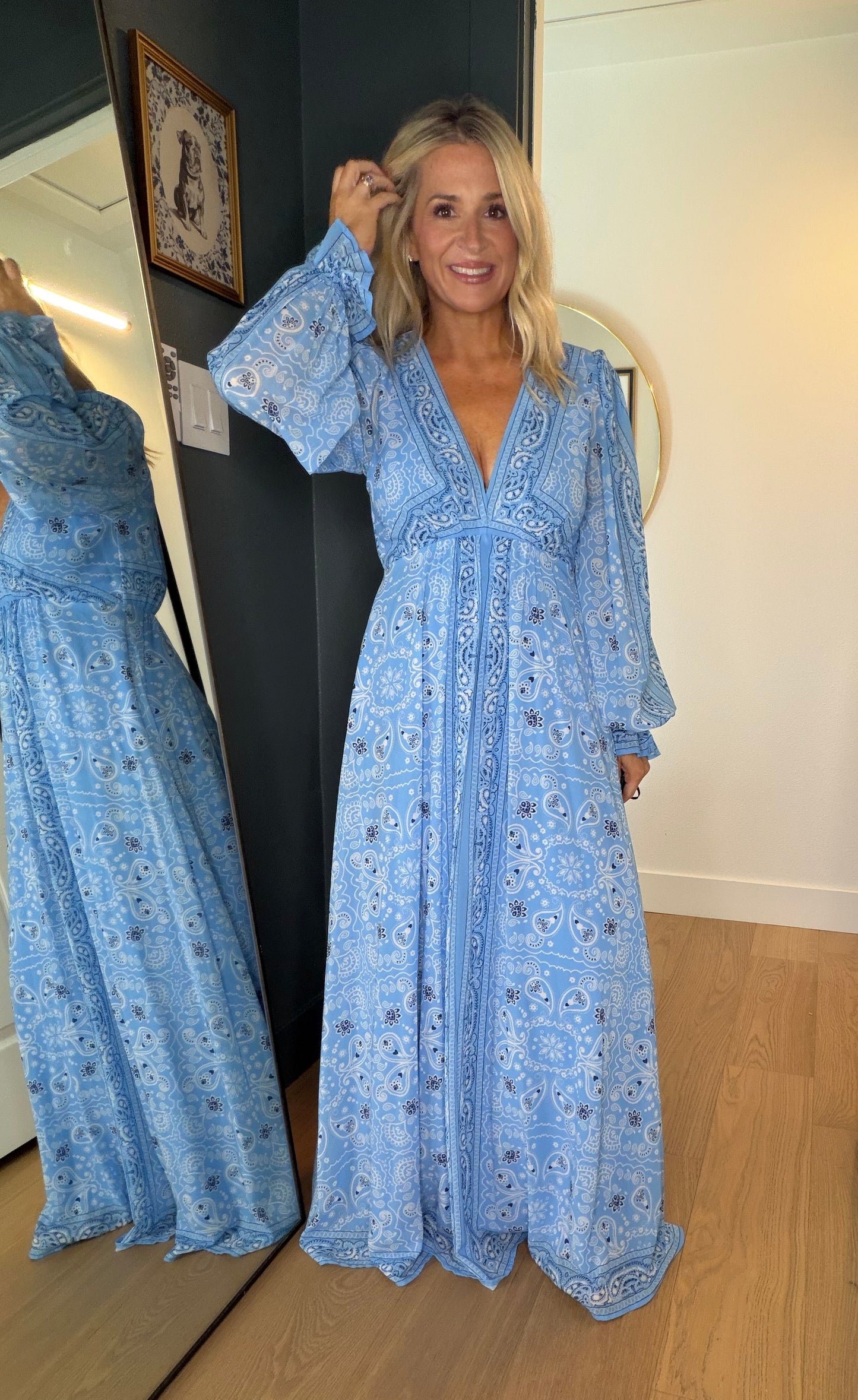 A woman wearing a light blue patterned maxi dress stands in a doorway, with her reflection visible in a mirror on the left. The dress has a bohemian-style floral print and long sleeves.