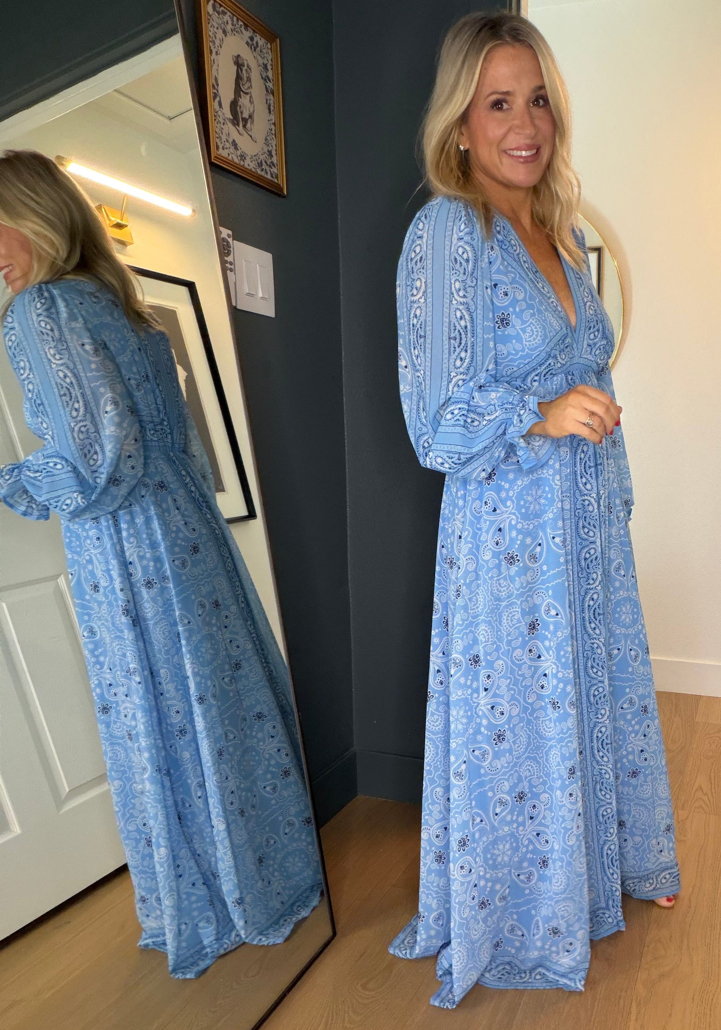 A woman wearing a light blue long-sleeve maxi dress with a white paisley pattern, standing in a doorway and looking at her reflection in a floor mirror.