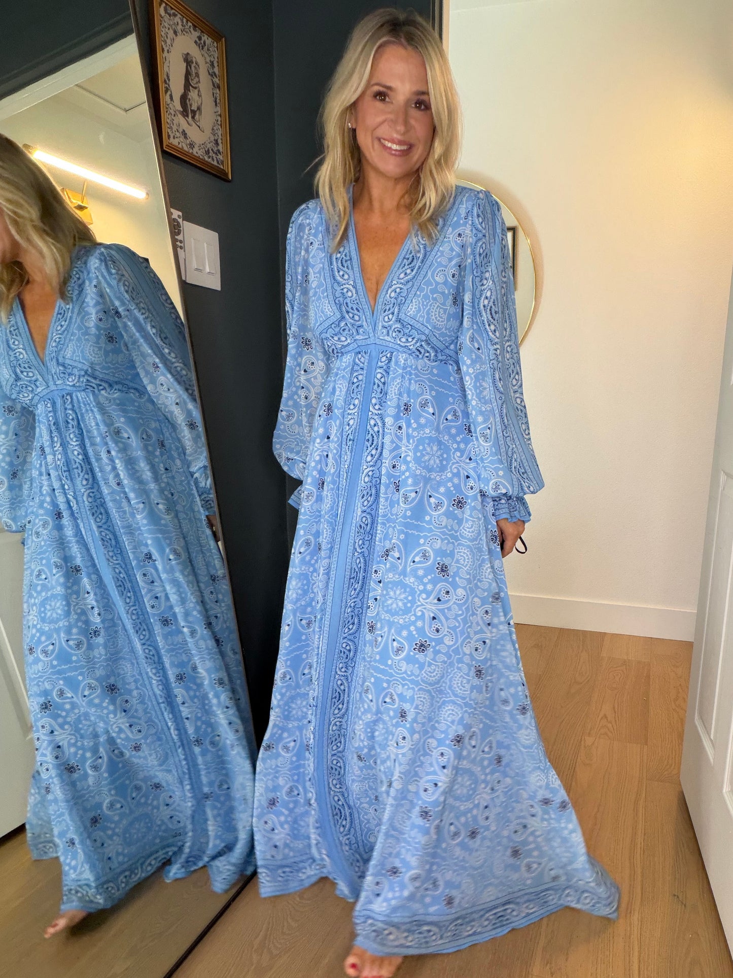 A woman wearing a light blue bohemian maxi dress with a floral print, standing in a doorway and looking at her reflection in a mirror.