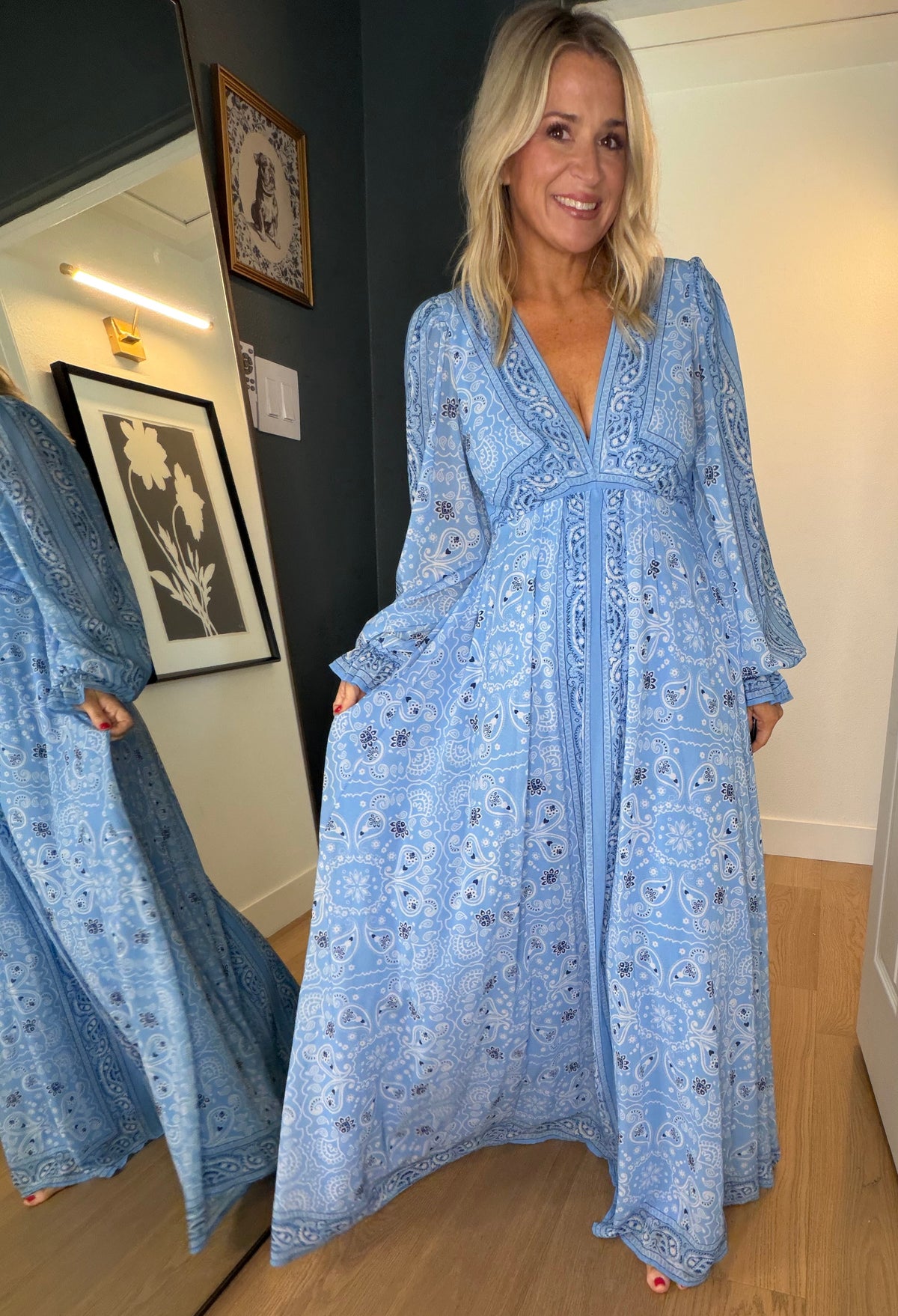 A woman wearing a light blue bohemian-style maxi dress with intricate embroidery, standing in a doorway of a home interior with wood floors and a mirror showing her side.
