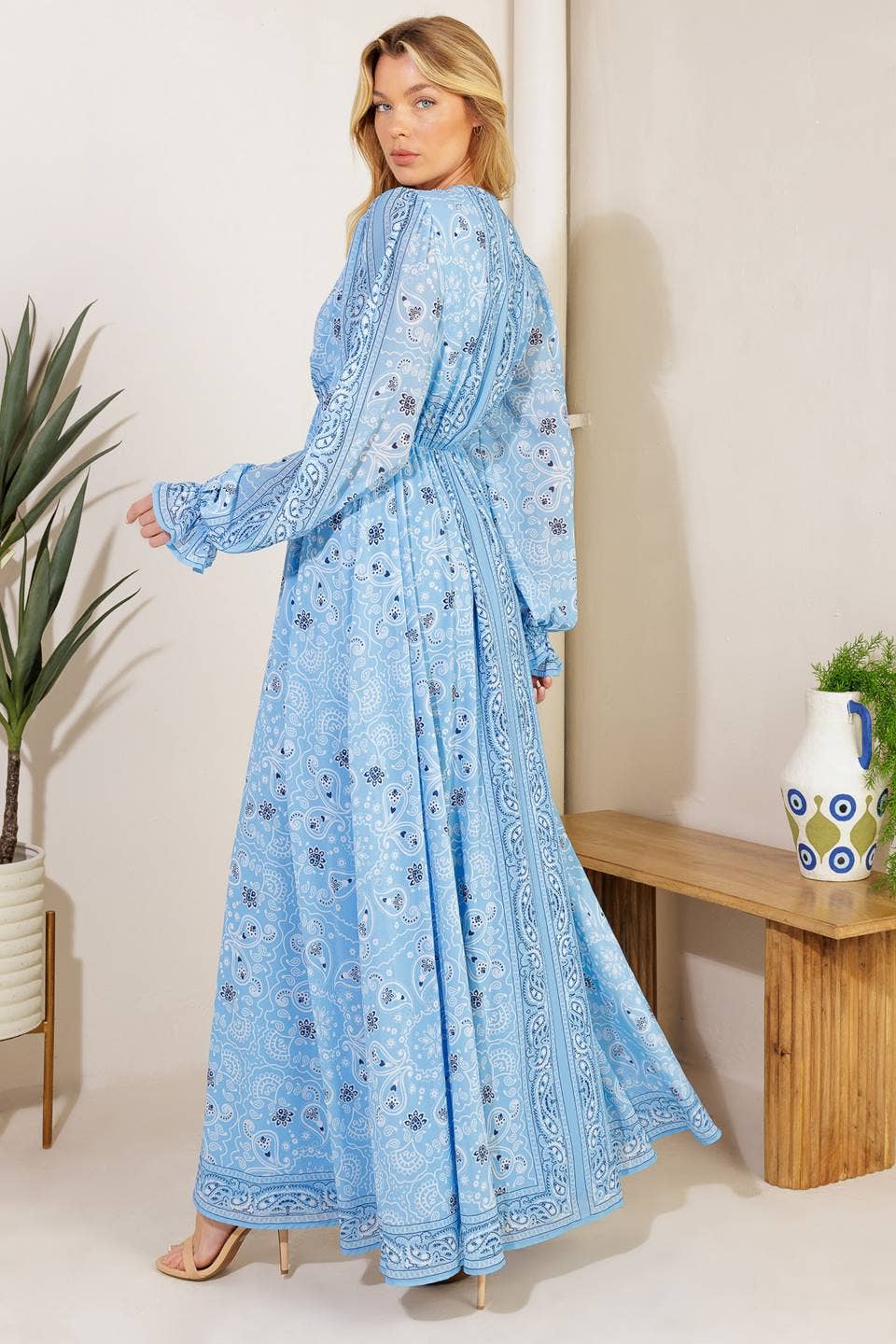 Woman wearing a blue patterned dress standing in a room with a plant and wooden bench.