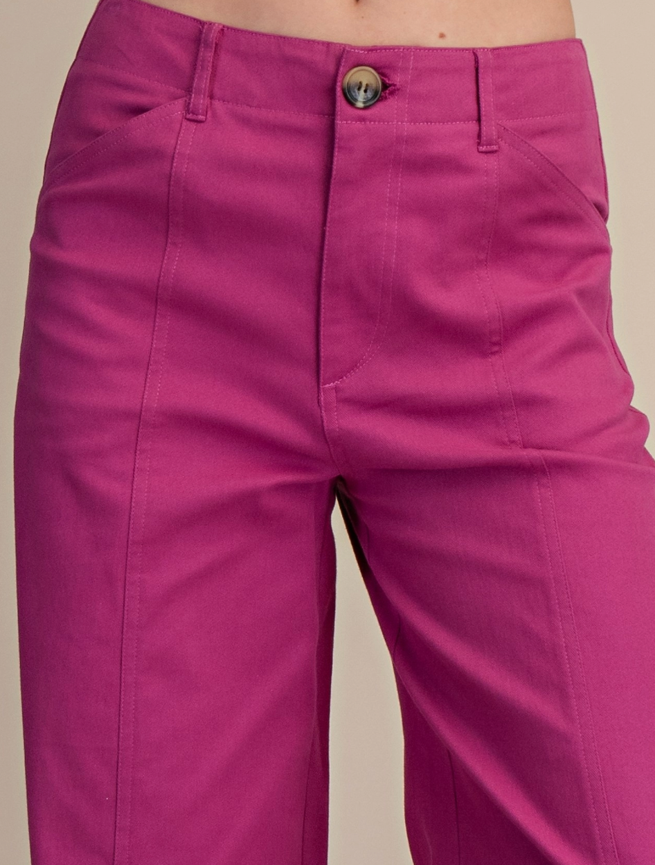 A close-up of a person wearing bright magenta pink trousers, showing the front waistband with a metallic button, belt loops, and front pockets against a plain beige background.
