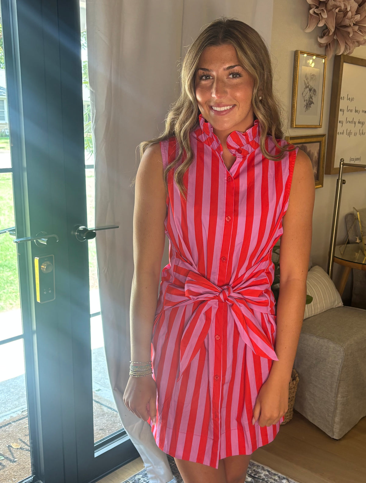 A woman wearing a sleeveless red and pink vertically striped shirt dress with a high collar and a self-tie waist, standing in a home interior near a glass door.