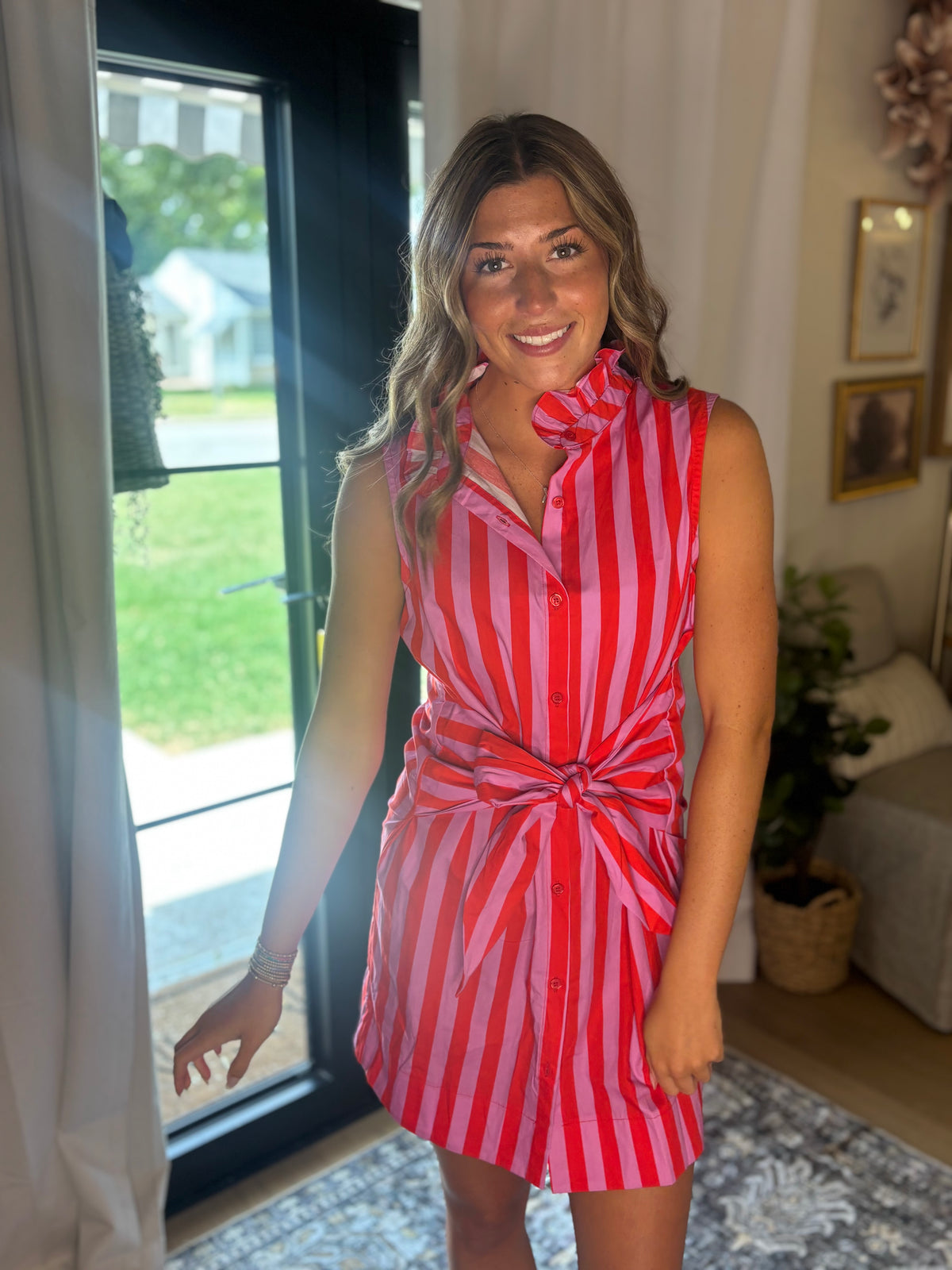 A woman wearing a sleeveless red and white vertically striped shirt dress with a tie waist, standing in a brightly lit room with a large window behind her.