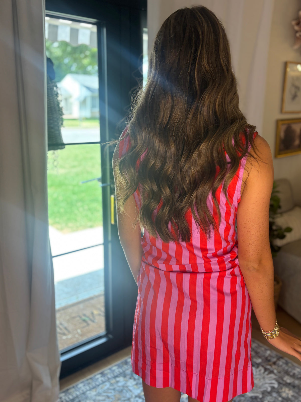 A woman viewed from behind wearing a sleeveless red and white vertically striped dress, standing in a home interior near a glass door.