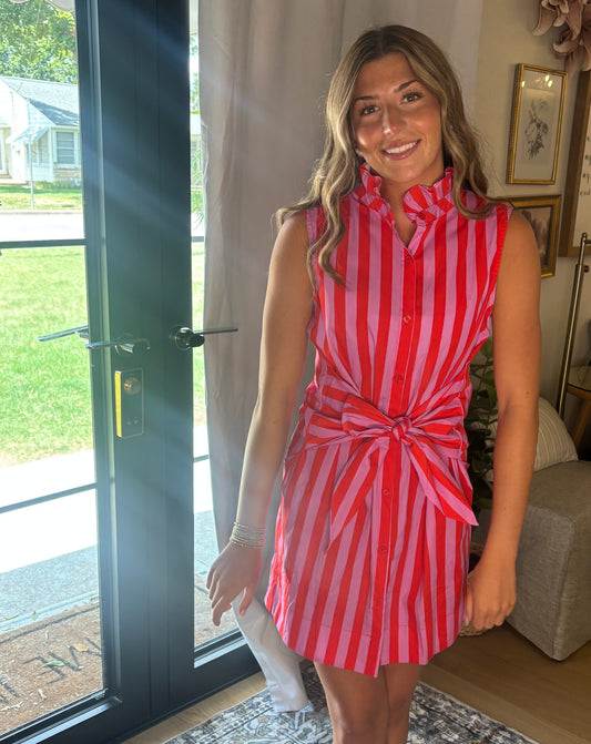 A woman wearing a sleeveless red and white vertically striped shirt dress with a tie waist, standing in a home interior near a glass door.