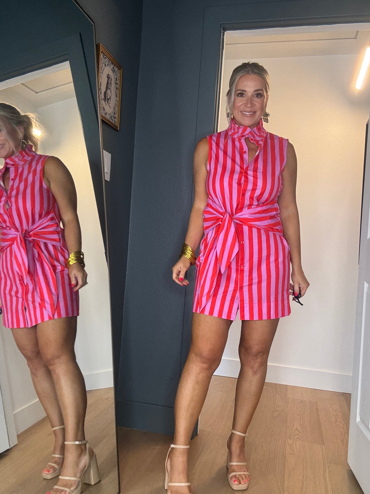 A woman modeling a sleeveless pink and red vertically striped mini dress with a tie waist and front buttons, standing in an indoor room with a mirror showing her side profile.