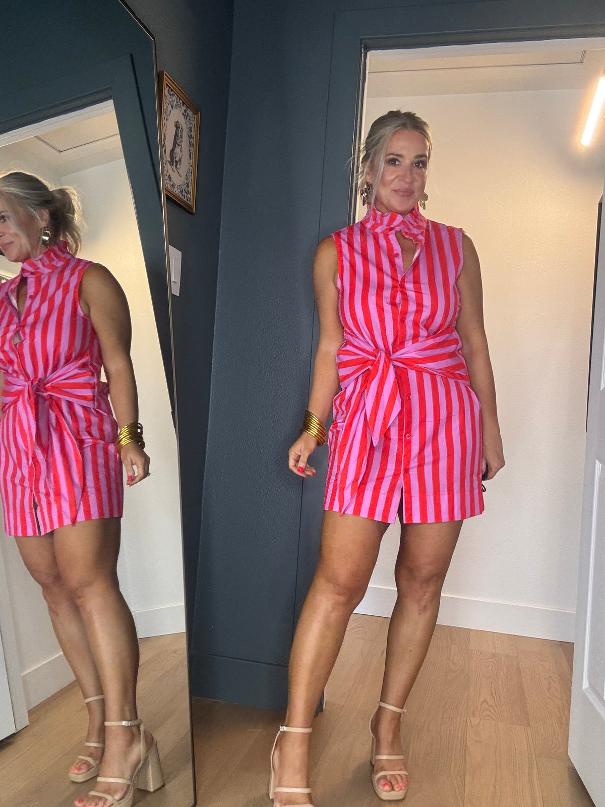 A woman wearing a pink and red striped sleeveless dress with a front twist detail, shown in a mirror selfie in an indoor room with wood flooring and a dark blue wall.