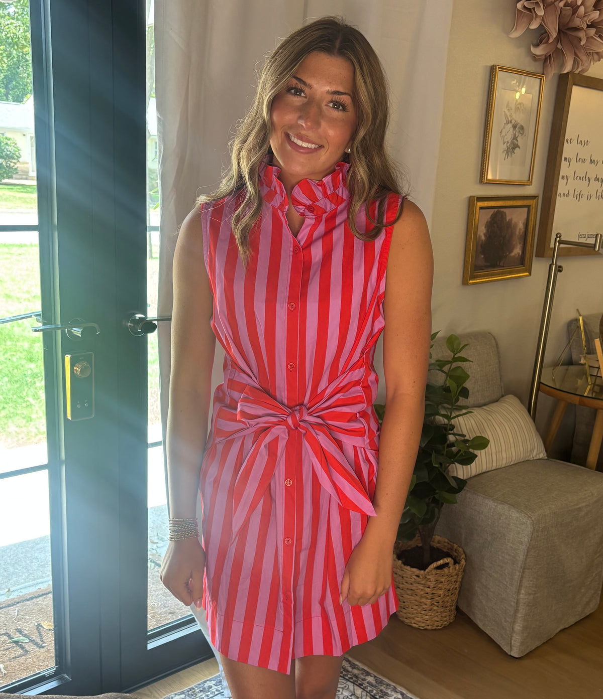 A woman wearing a sleeveless red and pink vertically striped shirt dress with a tie-neck and tie-waist detail, standing in a brightly lit room with a glass door, sofa, and framed art in the background.