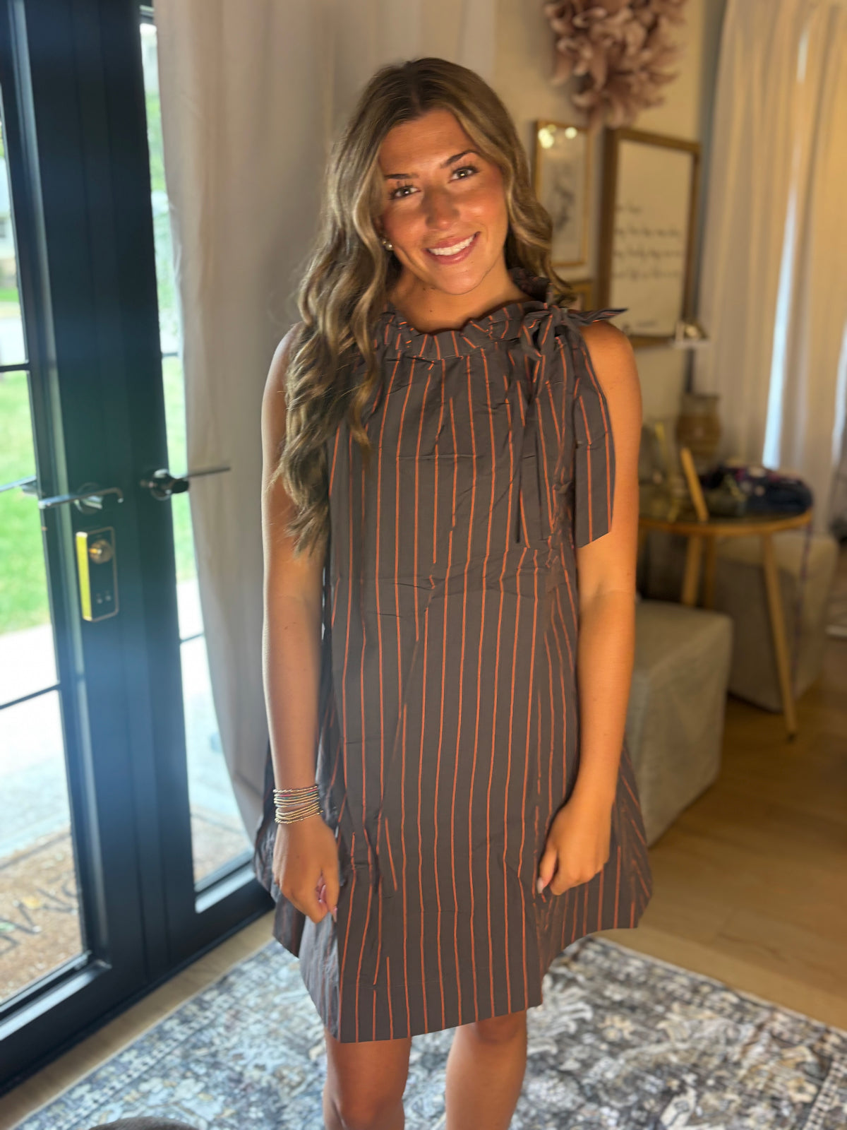 A woman modeling a sleeveless brown dress with thin orange vertical stripes, featuring a ruffled neckline and tie details on the shoulders, set in a home interior.