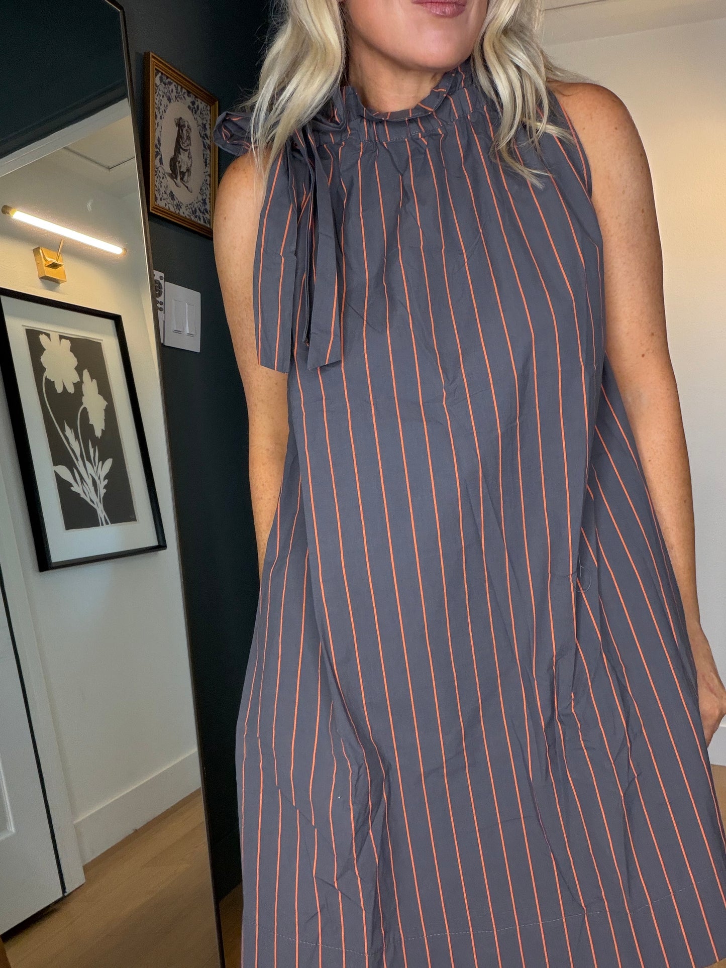 A woman wearing a sleeveless high-neck dress with vertical orange pinstripes on a dark gray background, standing indoors in a room with framed wall art.