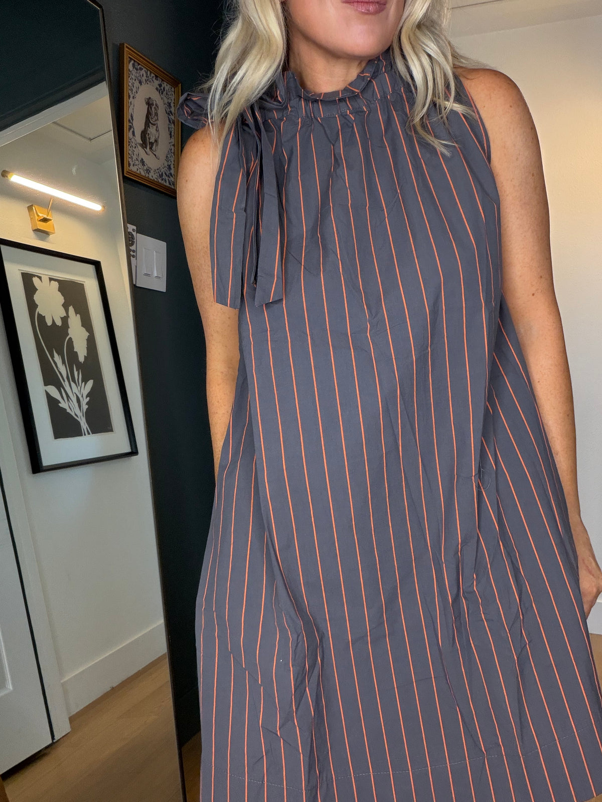 A woman wearing a sleeveless high-neck dress with vertical orange pinstripes on a dark gray background, standing indoors in a room with framed wall art.