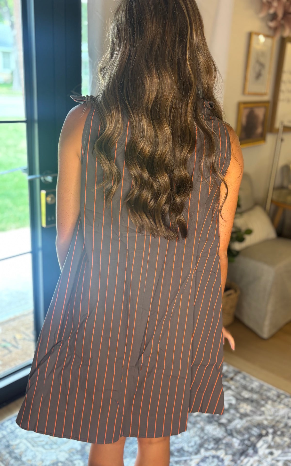 A woman viewed from behind wearing a sleeveless brown dress with thin orange vertical pinstripes, standing in a home interior near a glass door.
