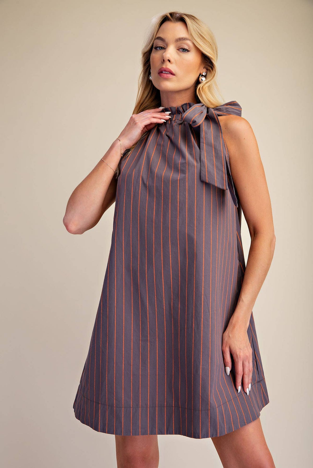A woman modeling a sleeveless high-neck dress with a bow at the collar, featuring vertical pinstripes in a muted purple-gray and orange-brown tone, against a plain beige studio background.