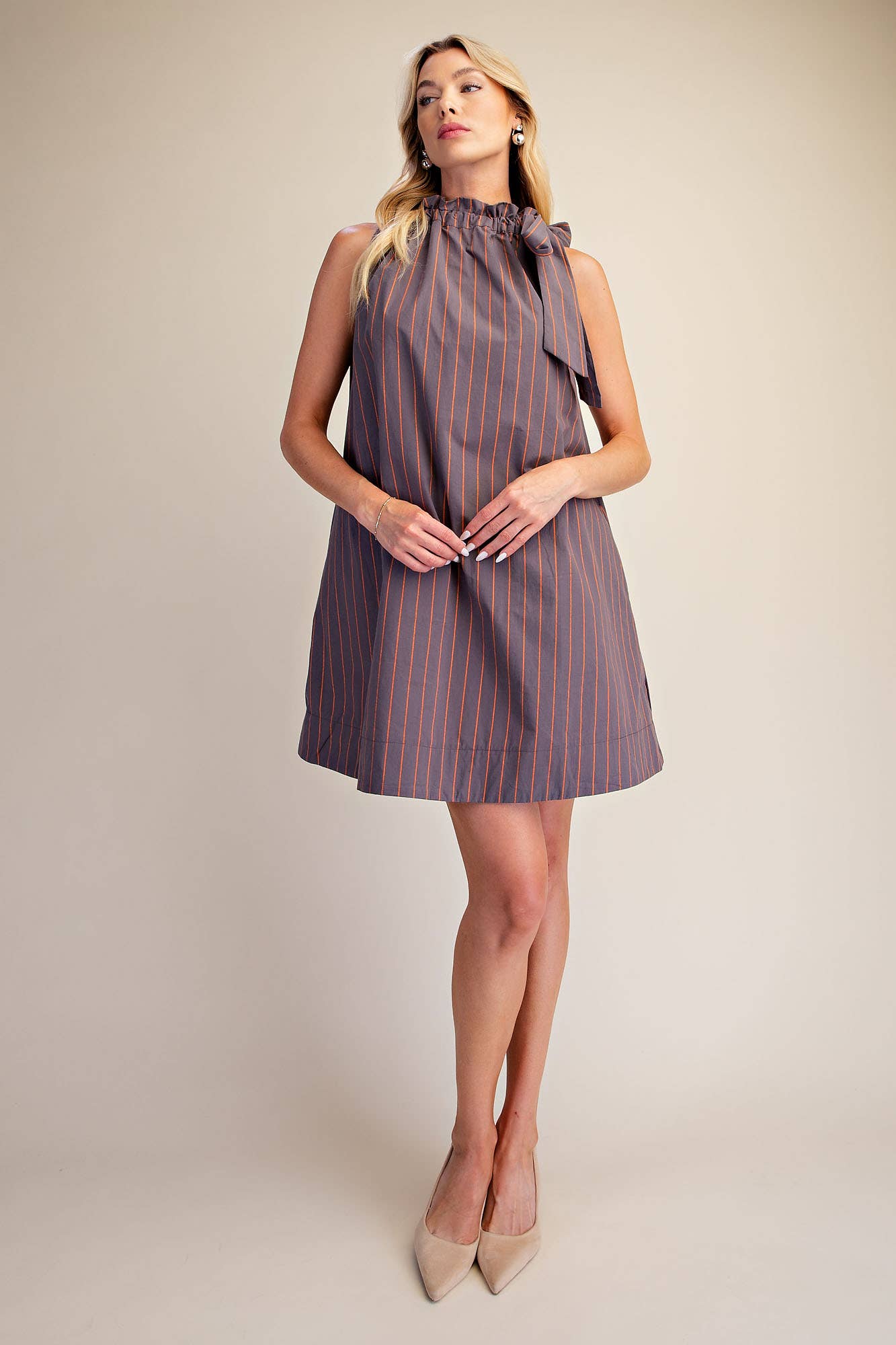 A woman modeling a sleeveless high-neck mini dress with vertical orange pinstripes on a muted grayish-purple base, paired with beige pointed-toe heels against a plain beige studio background.