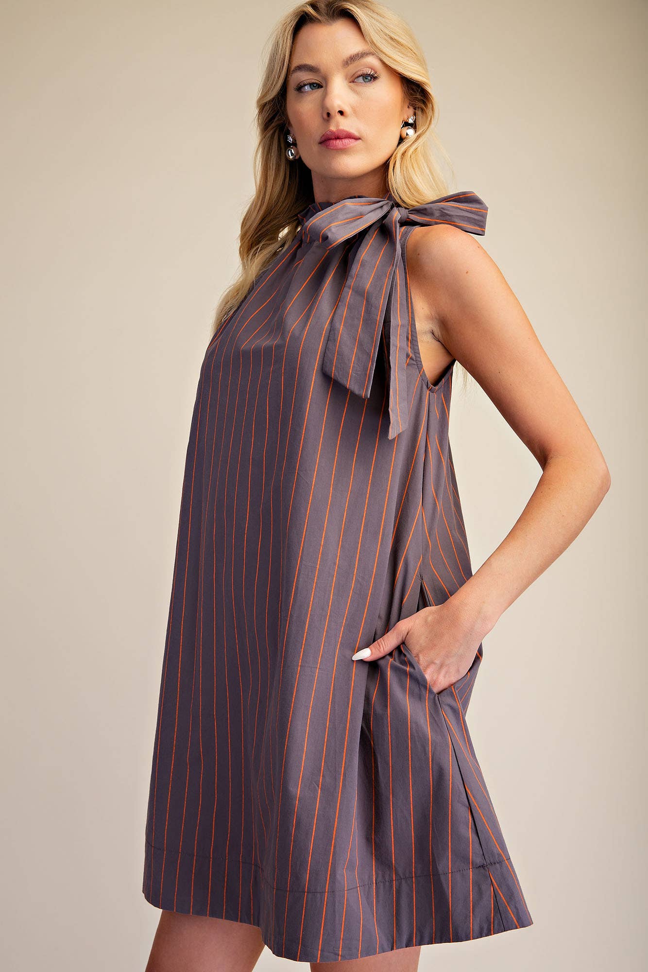 A woman modeling a sleeveless, knee-length dress with thin vertical orange pinstripes on a muted purple-gray base. The dress features a large bow detail at the high neckline and side pockets, photographed against a solid beige studio background.