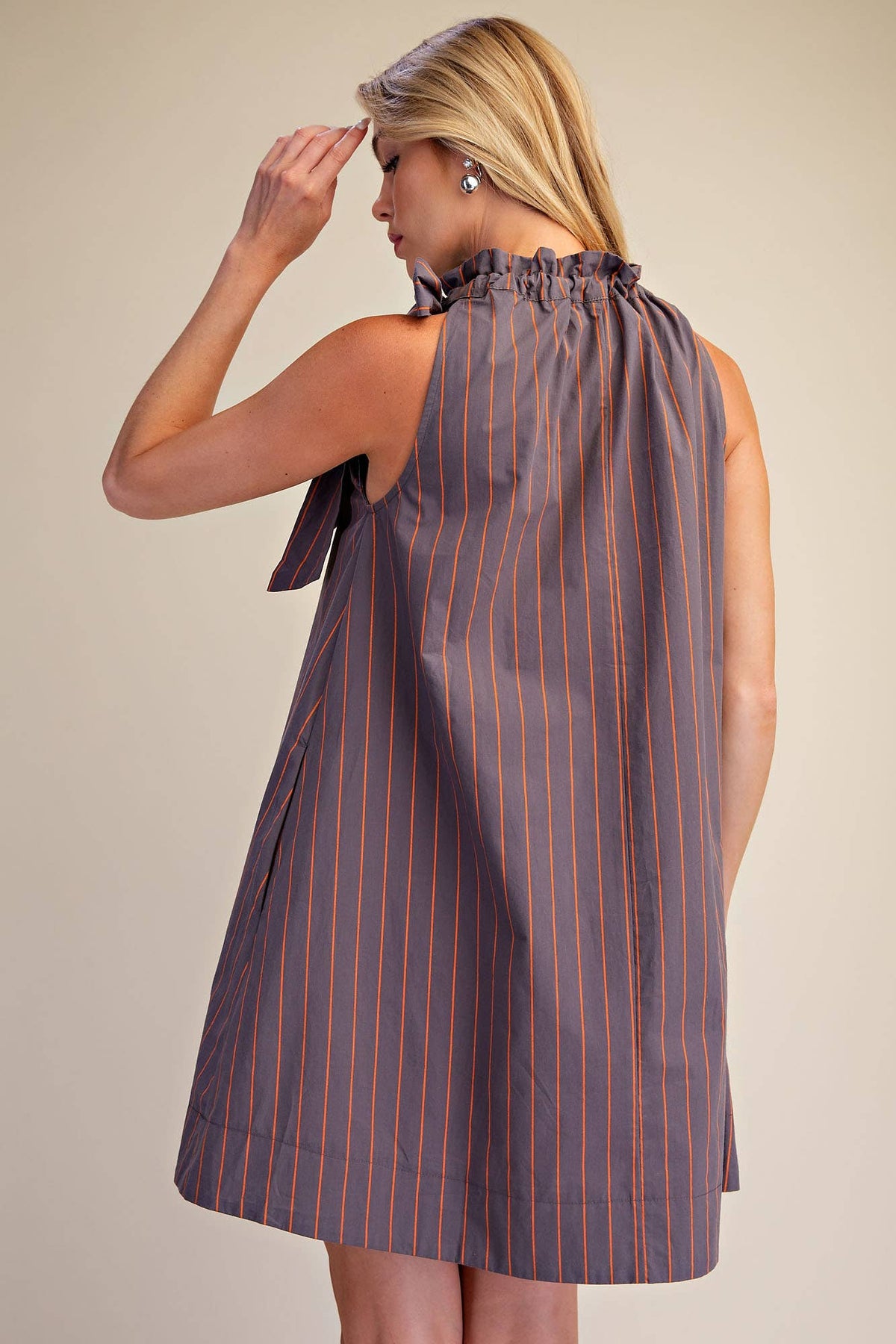A woman wearing a sleeveless, ruffled-neck dress with vertical orange pinstripes on a muted grayish-purple base, photographed against a plain beige studio background.