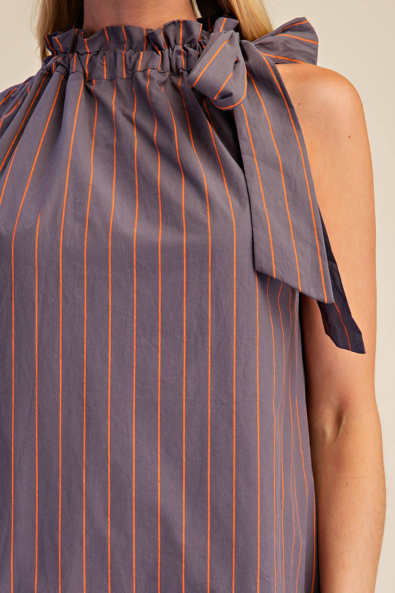 Close-up of a sleeveless top with a ruffled high neckline and side tie, featuring vertical orange pinstripes on a muted purple-gray fabric, worn by a model.