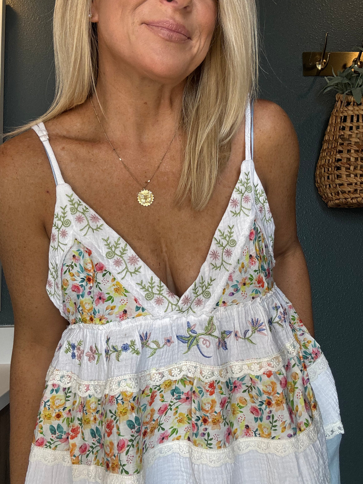 A close-up of a woman wearing a white spaghetti-strap dress with a colorful floral print and lace trim, paired with a gold pendant necklace.