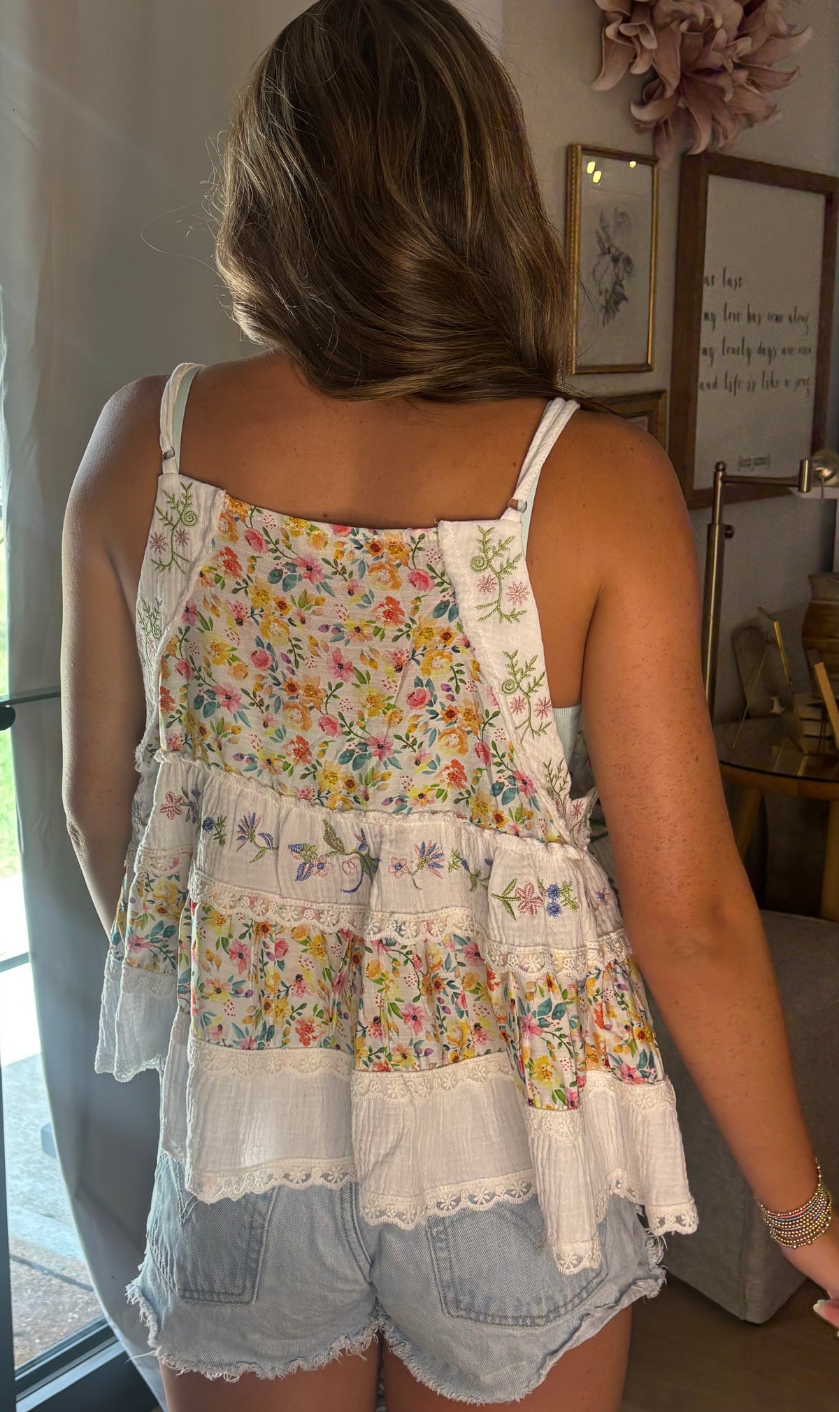 A woman shown from behind wearing a tiered spaghetti-strap top with a multicolor floral print and white lace trim, paired with denim shorts in an indoor setting.