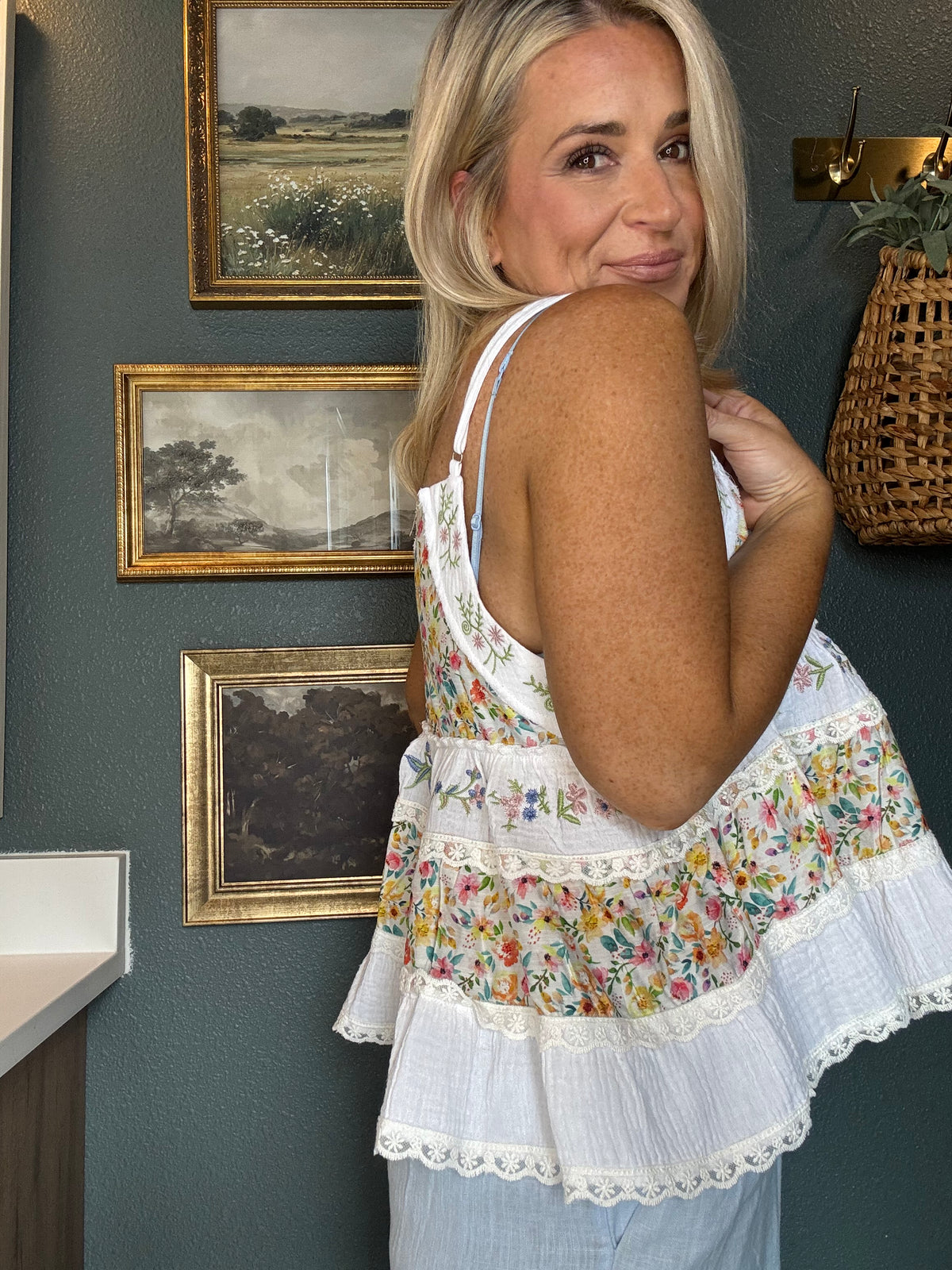 A woman wearing a white sleeveless ruffled blouse with a colorful floral print and lace trim, standing in front of a dark teal wall with framed landscape prints.