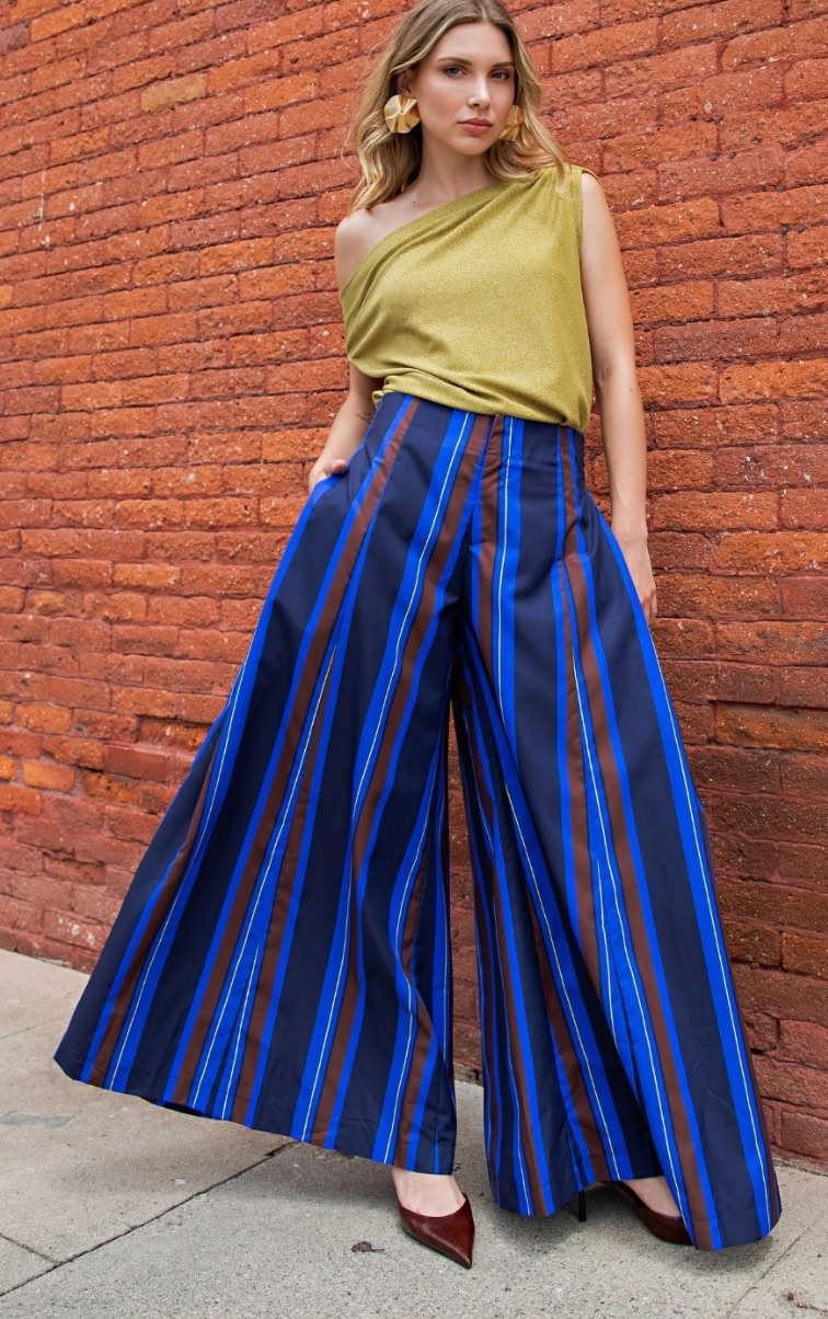A woman stands against a red brick wall wearing a one-shoulder olive green top and wide-leg trousers with vertical blue, navy, and rust-red stripes. She is wearing large gold earrings and dark red pointed shoes.