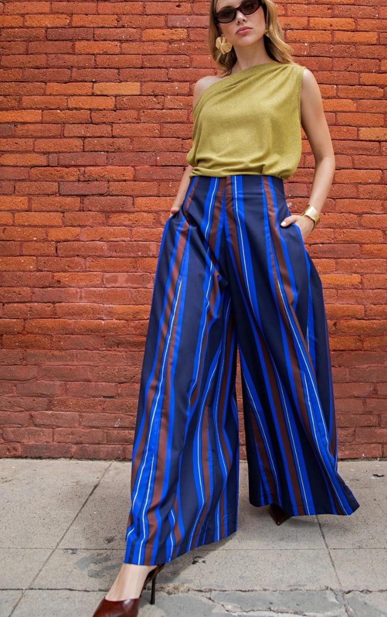 A woman models a stylish outfit consisting of a shimmering olive green one-shoulder top and wide-leg trousers with vertical blue, red, and navy stripes, standing against a red brick wall.