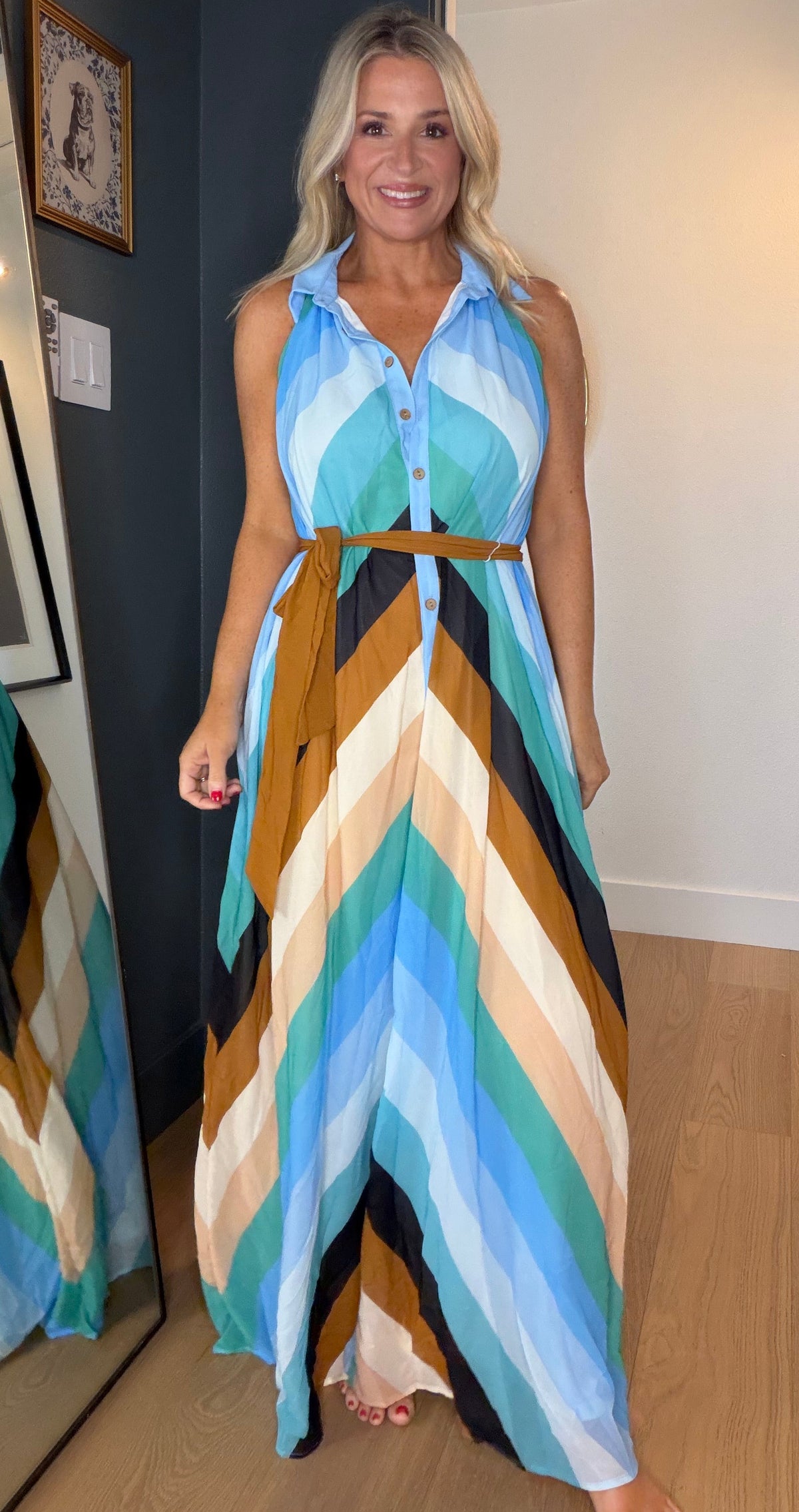 A woman modeling a sleeveless halter-neck maxi dress with a colorful chevron pattern in shades of blue, beige, brown, and white, standing in a hallway with a mirror showing her side profile.