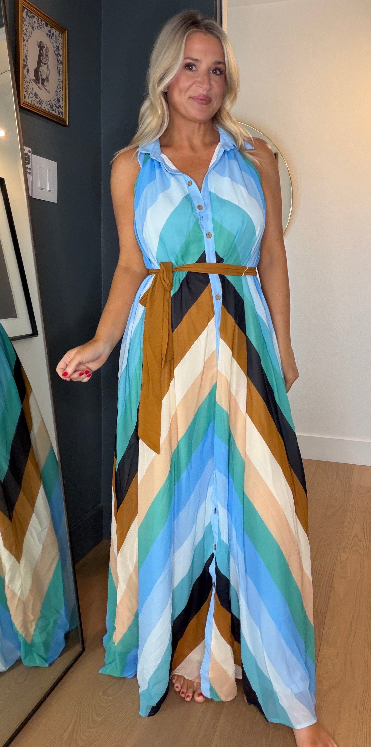 A woman wearing a sleeveless button-front maxi dress with a bold chevron pattern in light blue, teal, black, beige, and brown, standing in a doorway and looking at her reflection in a mirror.