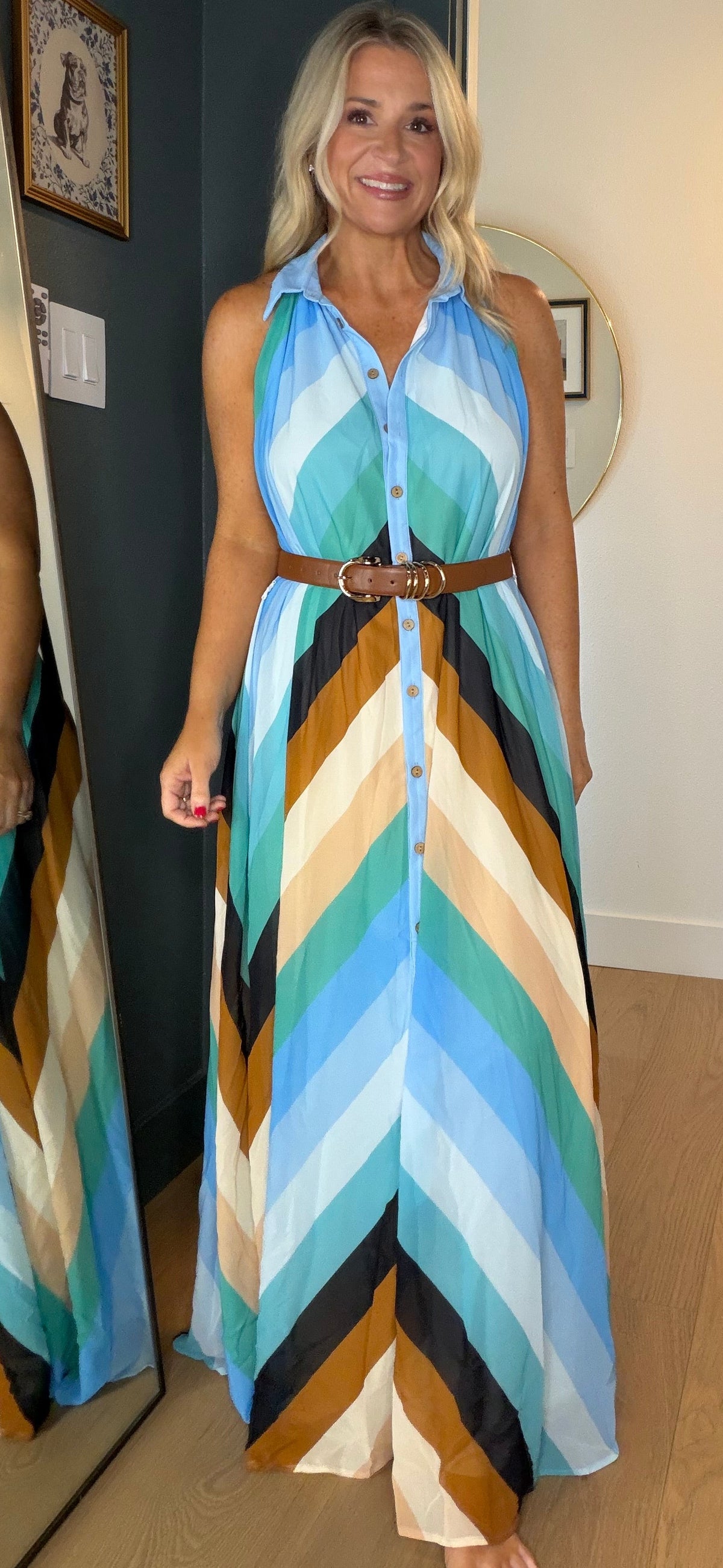 A woman wearing a sleeveless button-front maxi dress with a brown belt stands barefoot in a doorway, with her reflection visible in a large mirror on the left. The dress has a bold chevron pattern in shades of blue, beige, brown, black, and white.