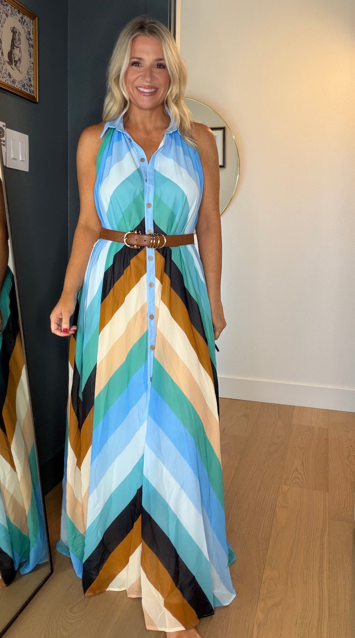A woman wearing a sleeveless halter maxi dress with a colorful chevron pattern in shades of blue, beige, brown, and black, styled with a brown belt. She is standing in a hallway with a wooden floor, a large mirror showing her reflection, and white doors on both sides.