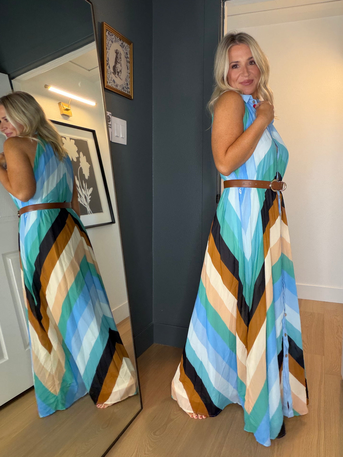 A woman stands in a doorway wearing a sleeveless halter maxi dress with a bold multicolor chevron pattern in shades of blue, teal, brown, cream, and black, tied at the waist with a thin brown belt. A large mirror on the left reflects her side profile of the dress.