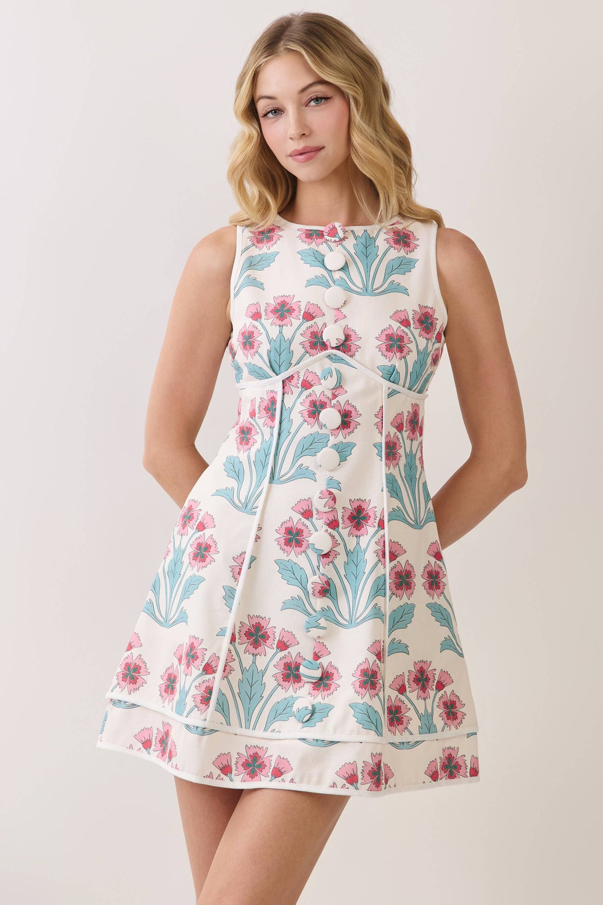 A sleeveless fit-and-flare dress with a pink and teal floral print on a cream background, modeled against a light beige studio backdrop.