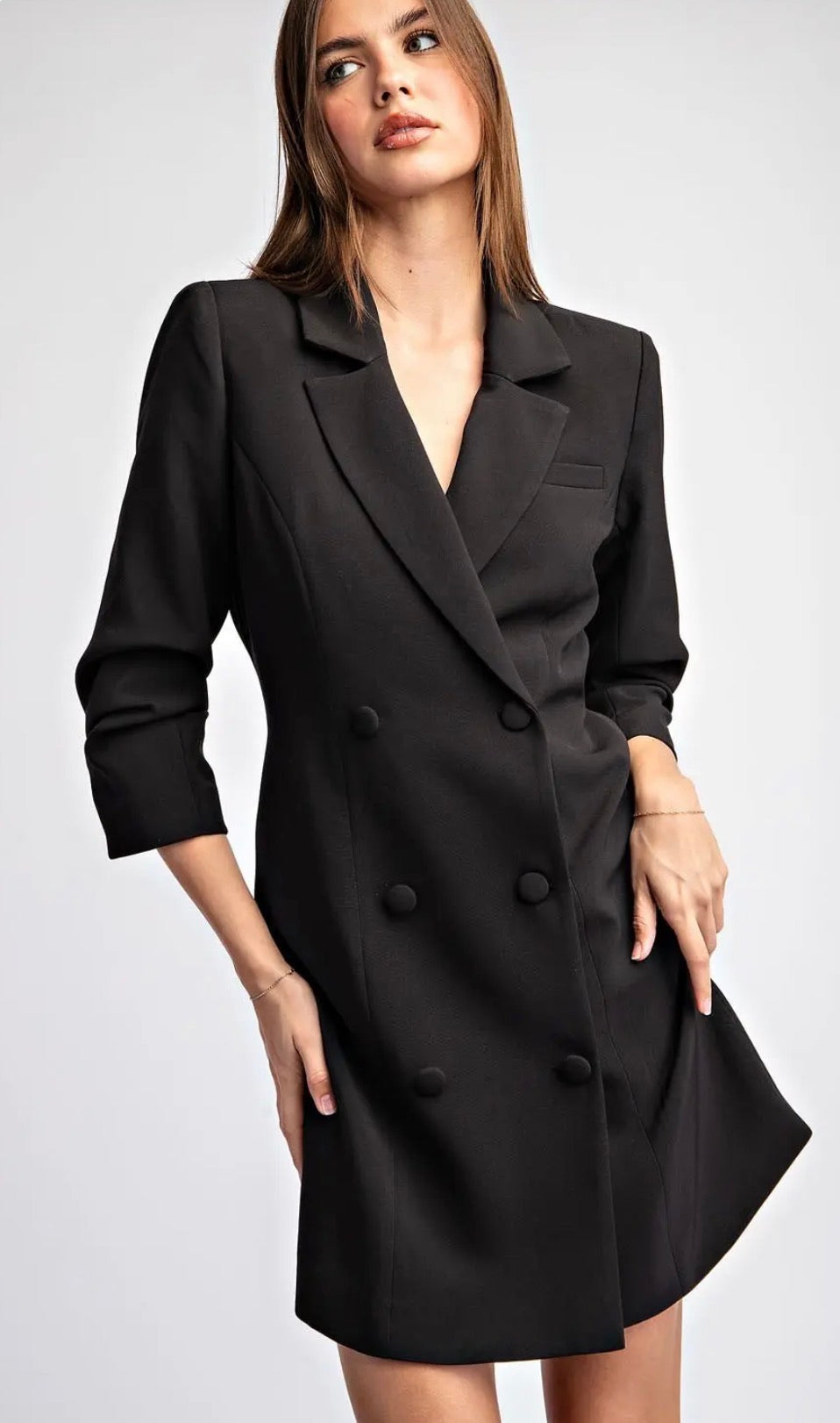 A woman modeling a black double-breasted blazer dress with wide lapels and three-quarter sleeves against a light gray studio background.