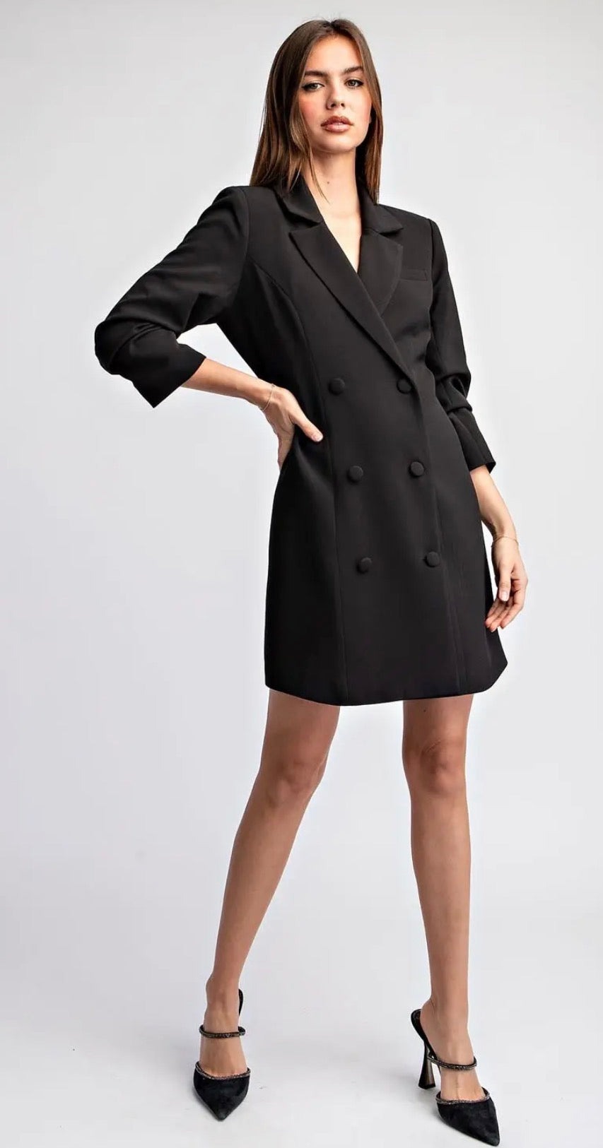 A woman modeling a black double-breasted blazer dress with three-quarter sleeves, paired with black pointed-toe heels, against a light gray studio background.