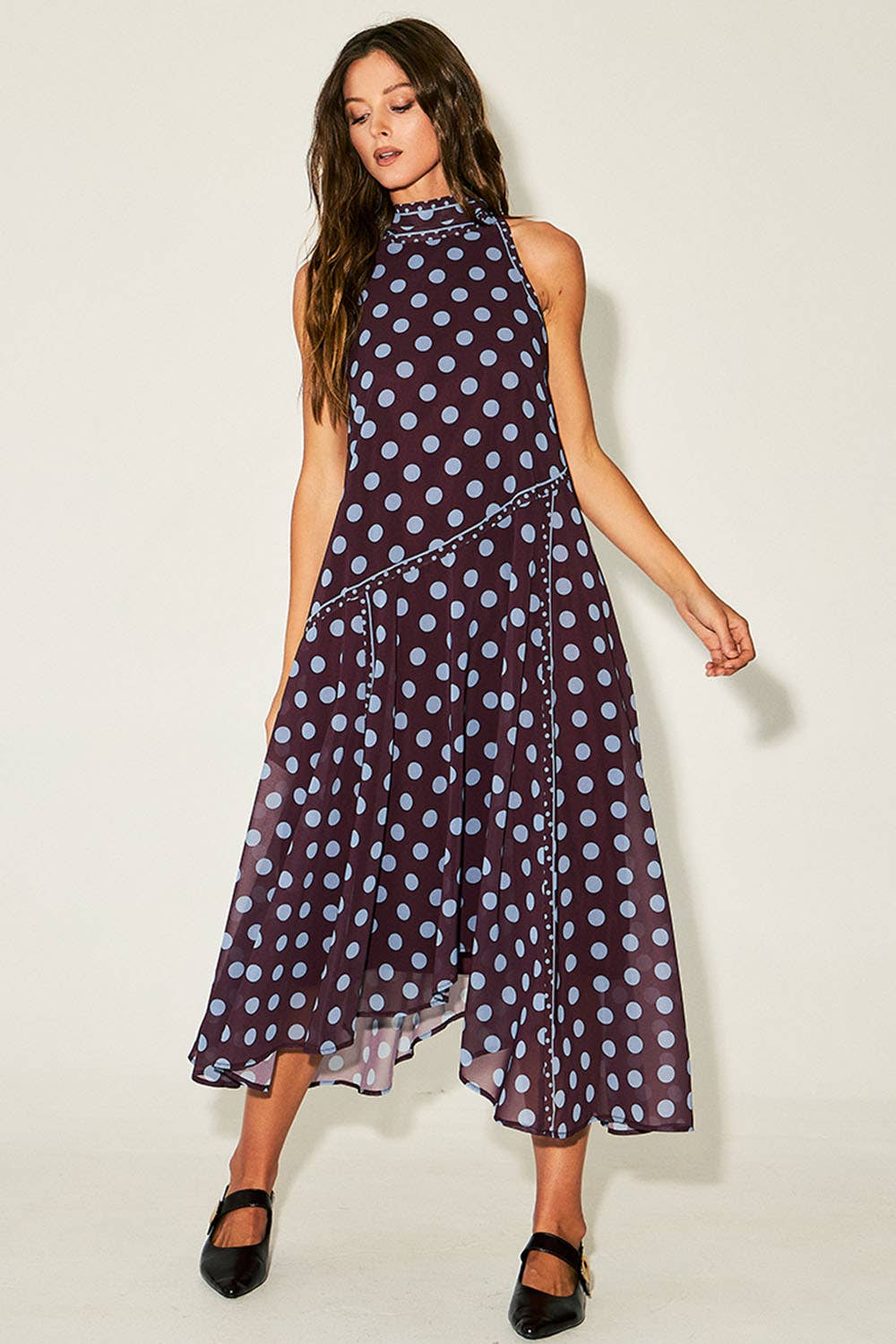 A woman modeling a sleeveless high-neck midi dress with a dark purple base and light blue polka dots, paired with black Mary Jane shoes, against a plain off-white studio background.