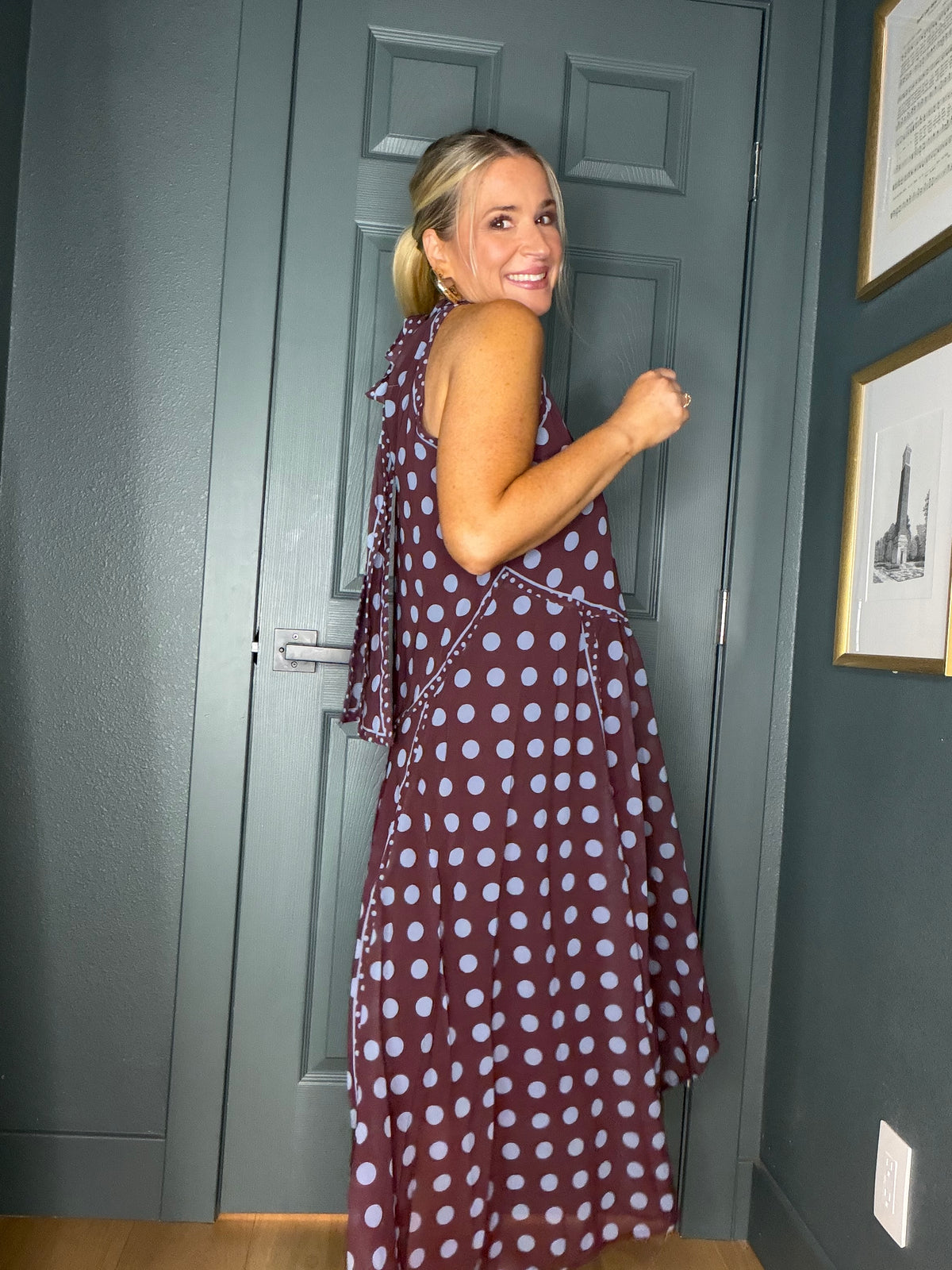 A woman wearing a sleeveless, one-shoulder maxi dress with a burgundy base and light blue polka dots, posing in front of a dark gray paneled door.