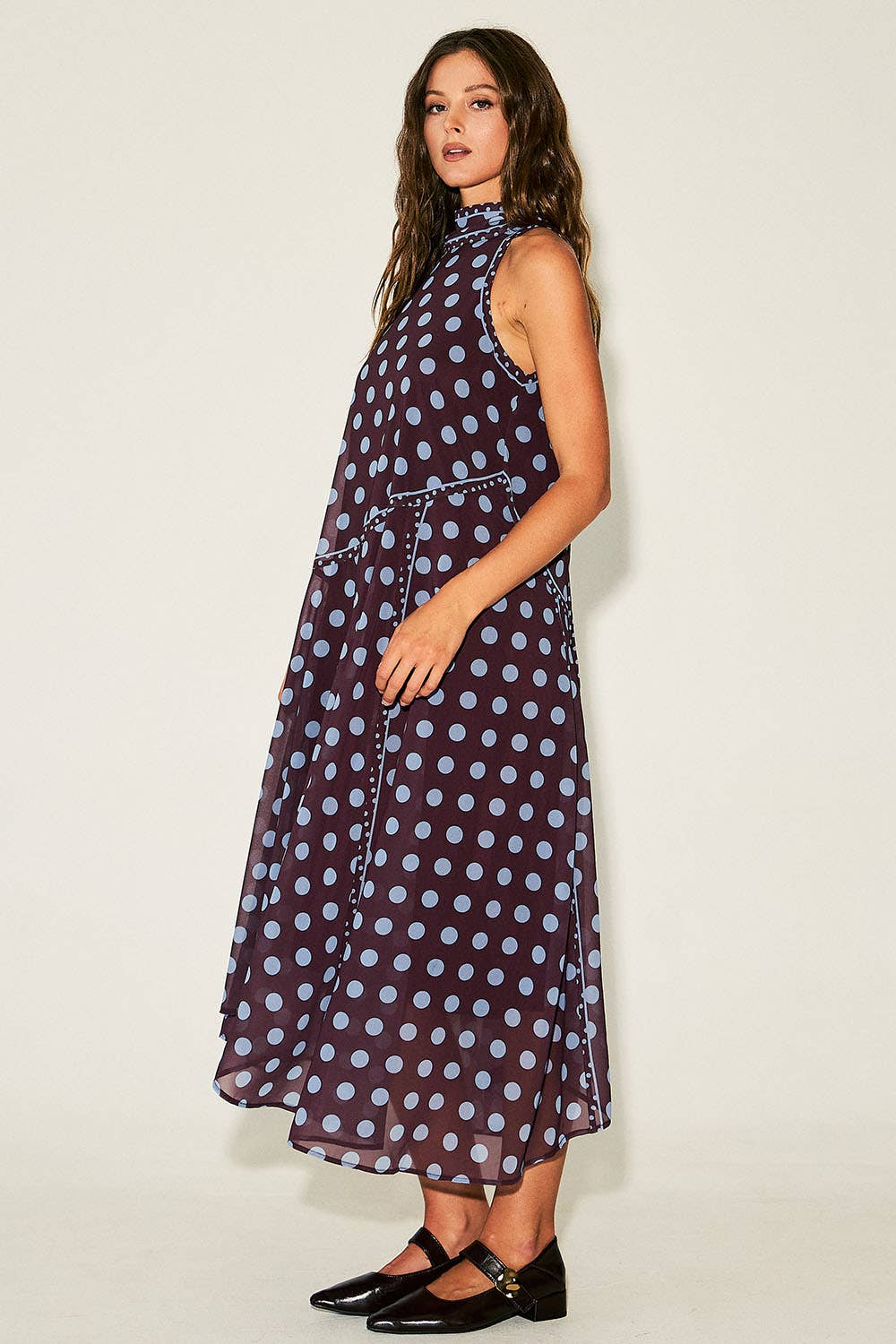 A woman modeling a sleeveless high-neck midi dress with a dark purple base and light blue polka dots, paired with black Mary Jane shoes, against a plain off-white studio background.