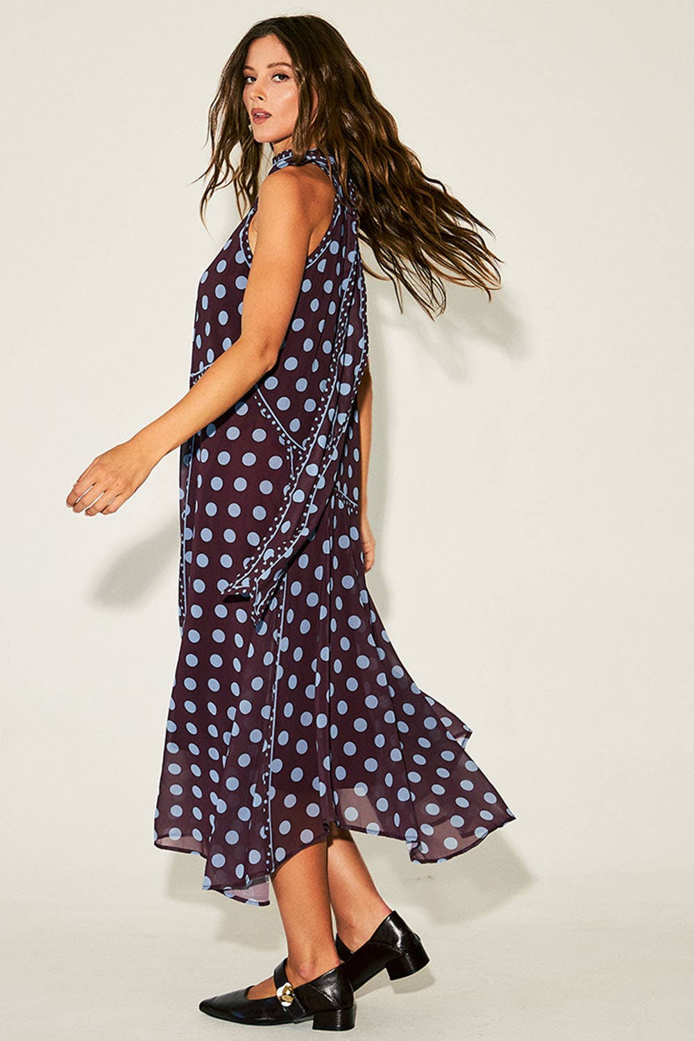 A woman modeling a sleeveless, high-neck midi dress with a dark purple base and light blue polka dots, paired with black Mary Jane heels, against a plain off-white studio background.